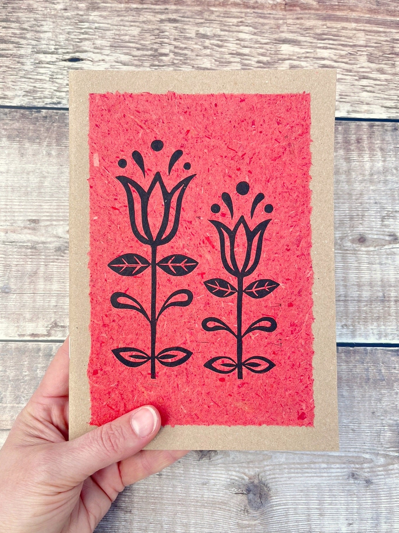 A5 kraft sketchbook with an original lino print of Tulips on handmade brick red paper. Held in a hand against a wooden background.