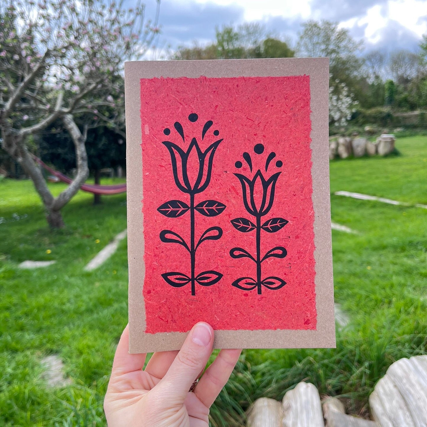 A5 kraft sketchbook with an original lino print of Tulips on handmade brick red paper. Held in a hand in a garden.