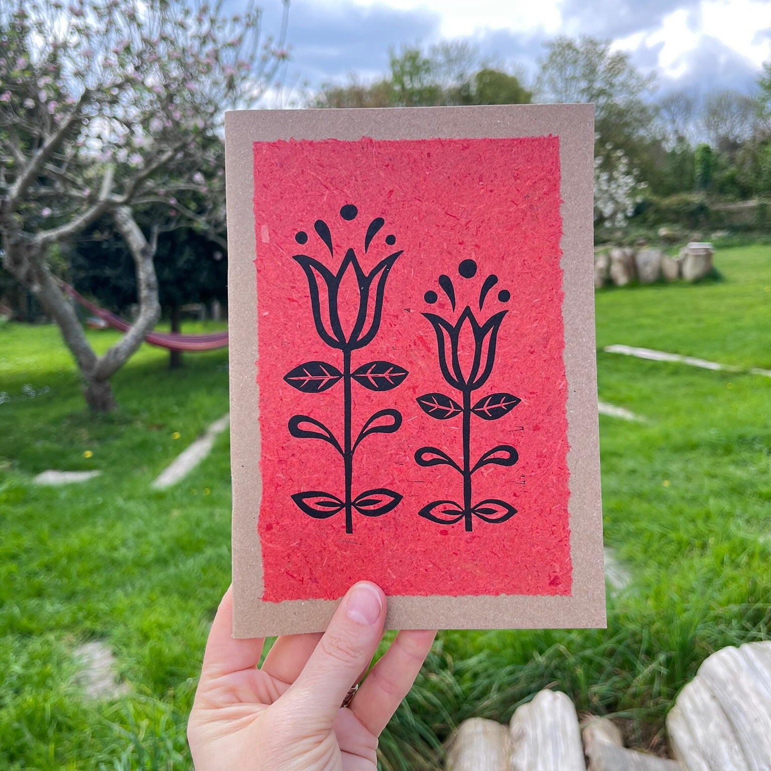 A5 kraft sketchbook with an original lino print of Tulips on handmade brick red paper. Held in a hand in a garden.