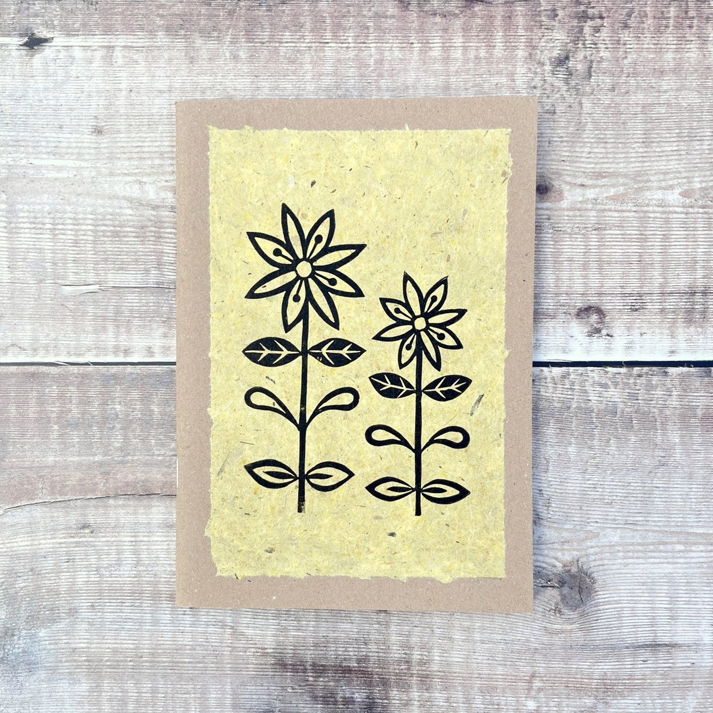 A5 kraft sketchbook with an original lino print of daisies on handmade yellow paper. Laid on a wooden background.