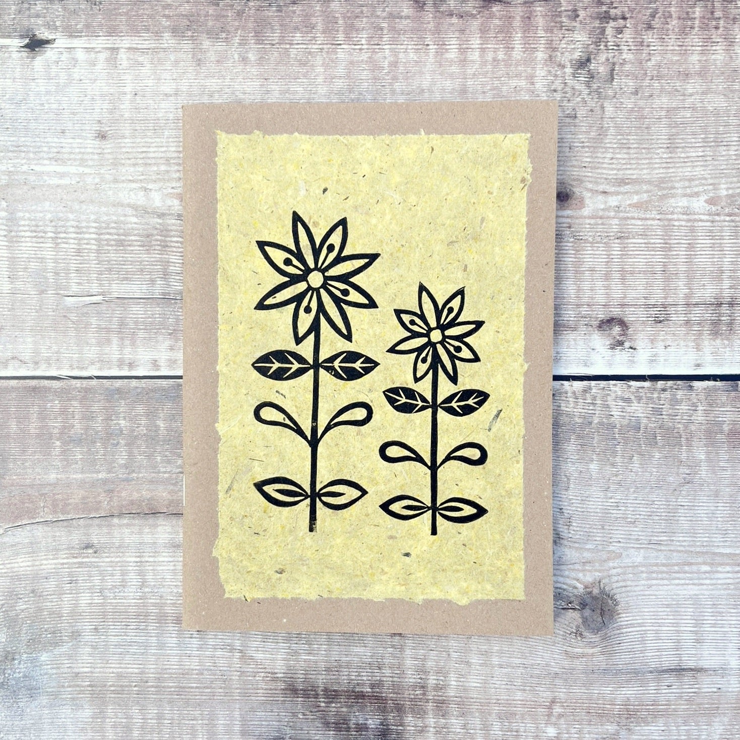 A5 kraft sketchbook with an original lino print of daisies on handmade yellow paper. Laid on a wooden background.