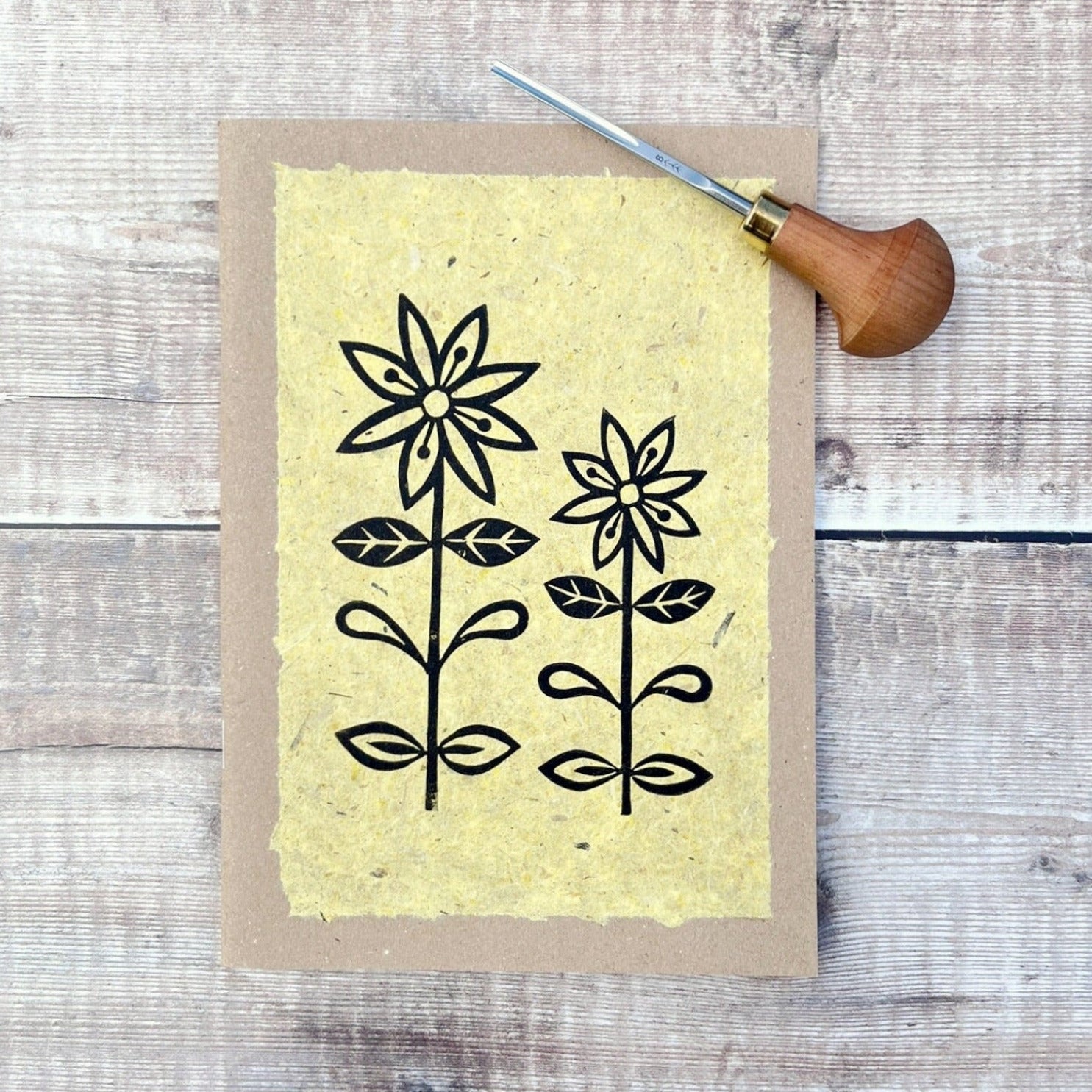 A5 kraft sketchbook with an original lino print of daisies on handmade yellow paper. Photo includes lino cutting tool laid on a wooden background.