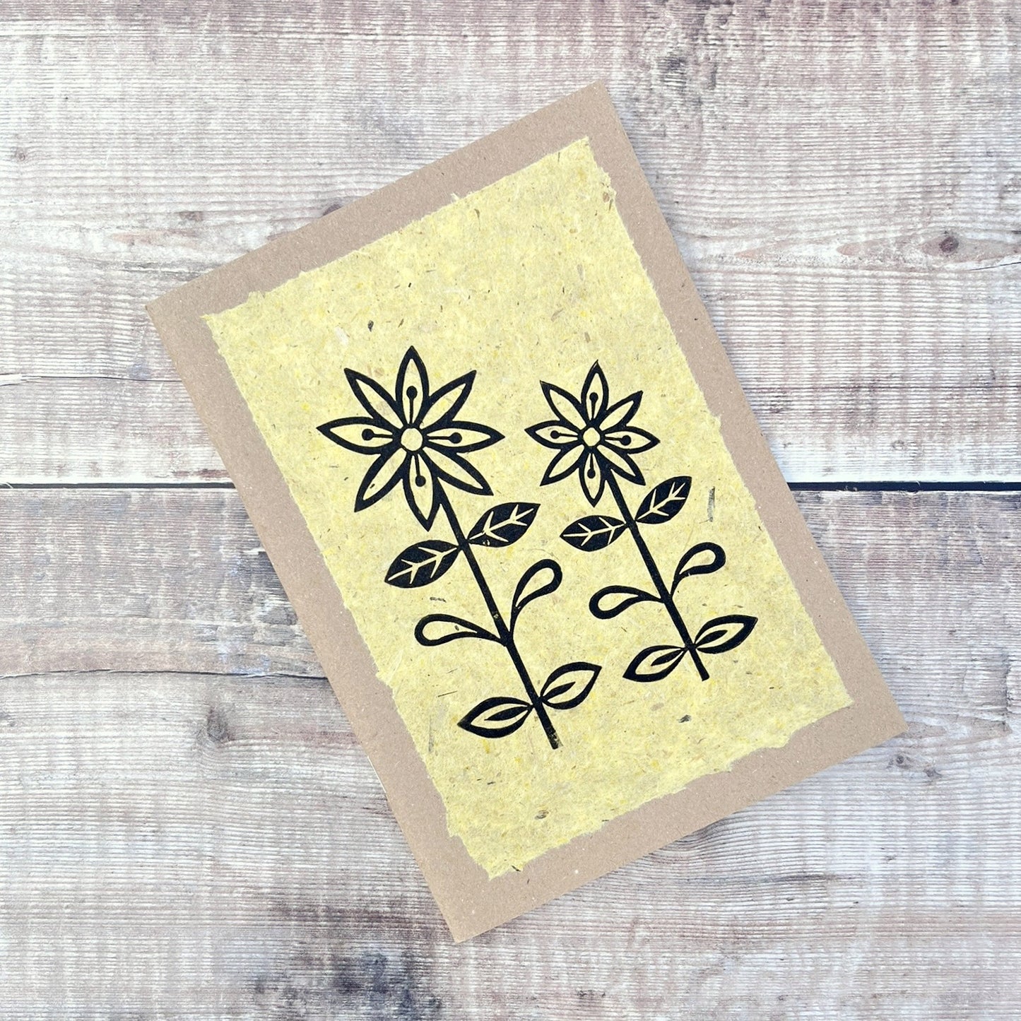 A5 kraft sketchbook with an original lino print of daisies on handmade yellow paper. Laid on a wooden background.