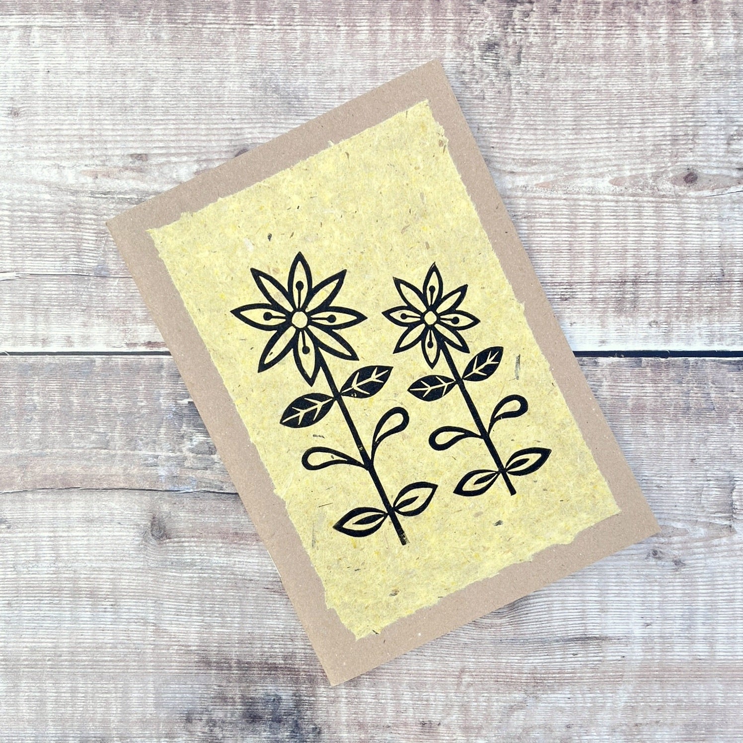 A5 kraft sketchbook with an original lino print of daisies on handmade yellow paper. Laid on a wooden background.