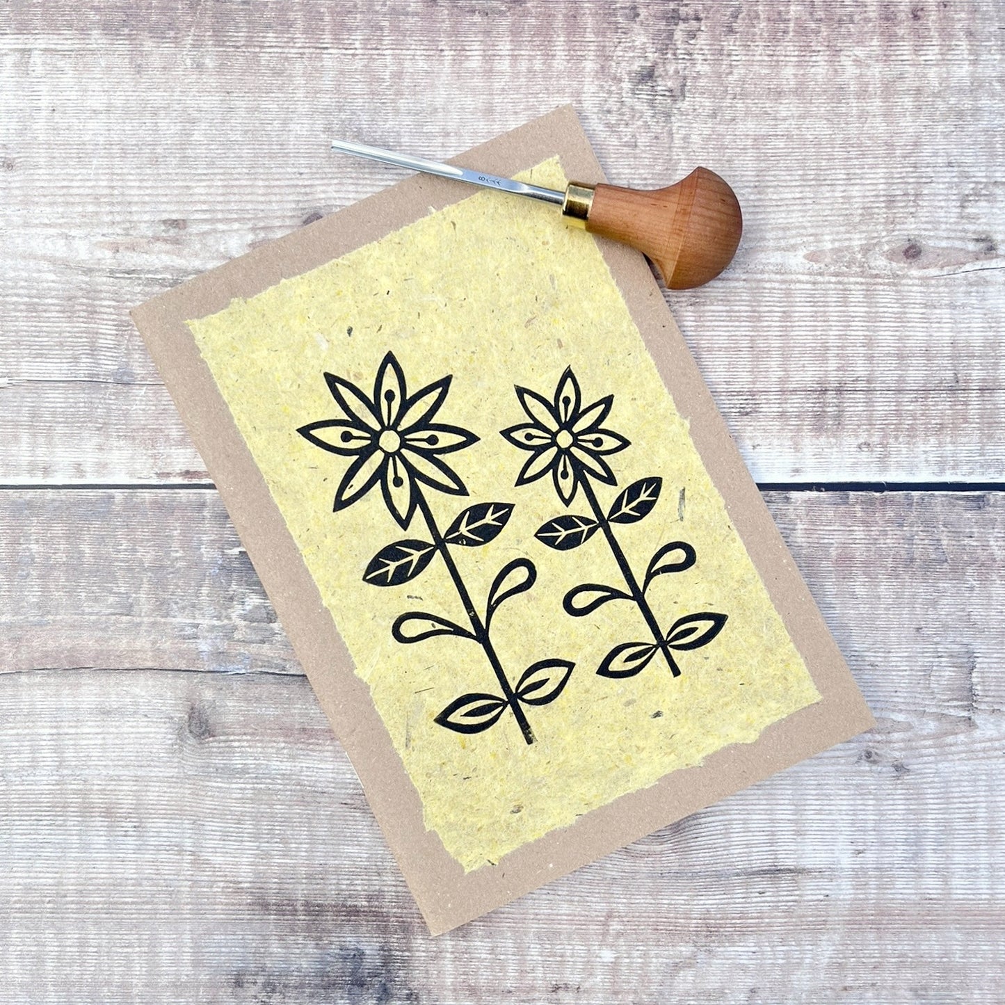 A5 kraft sketchbook with an original lino print of daisies on handmade yellow paper. Photo includes lino cutting tool laid on a wooden background.
