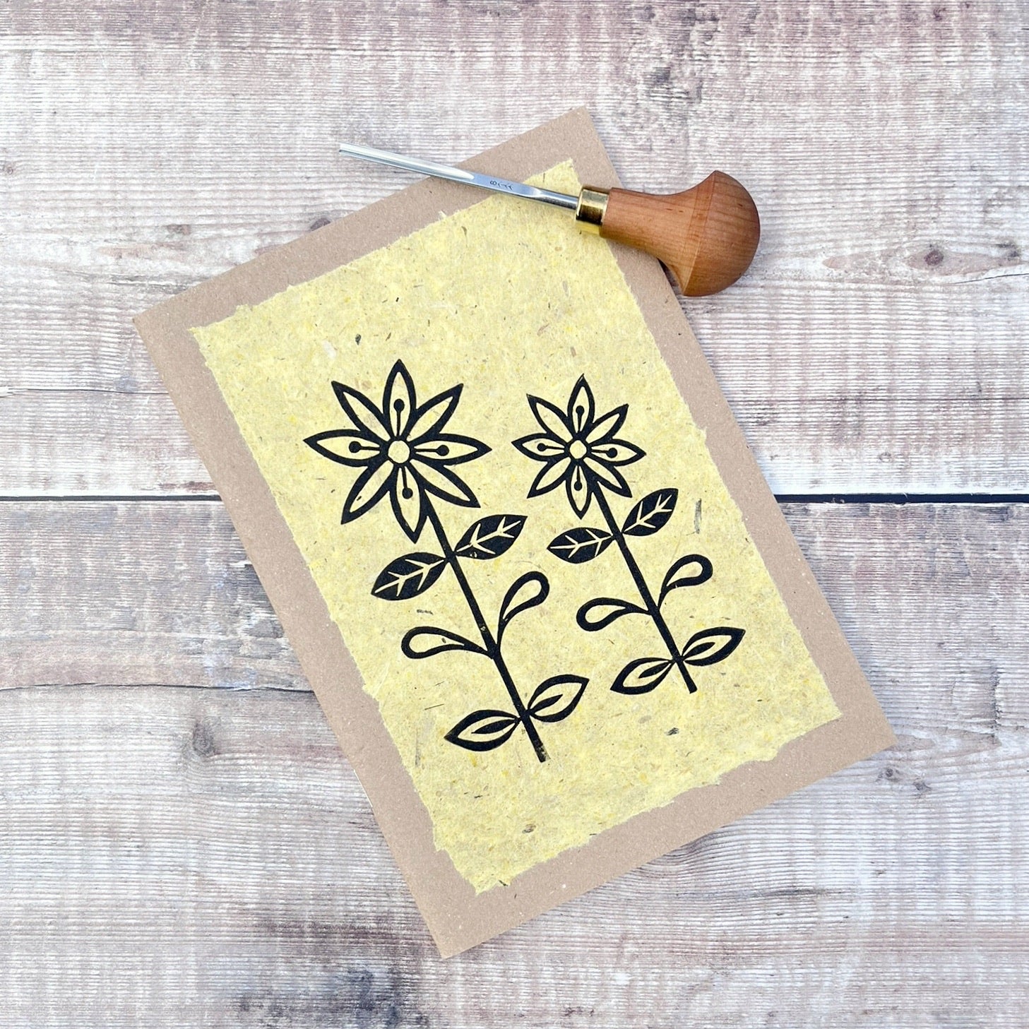 A5 kraft sketchbook with an original lino print of daisies on handmade yellow paper. Photo includes lino cutting tool laid on a wooden background.