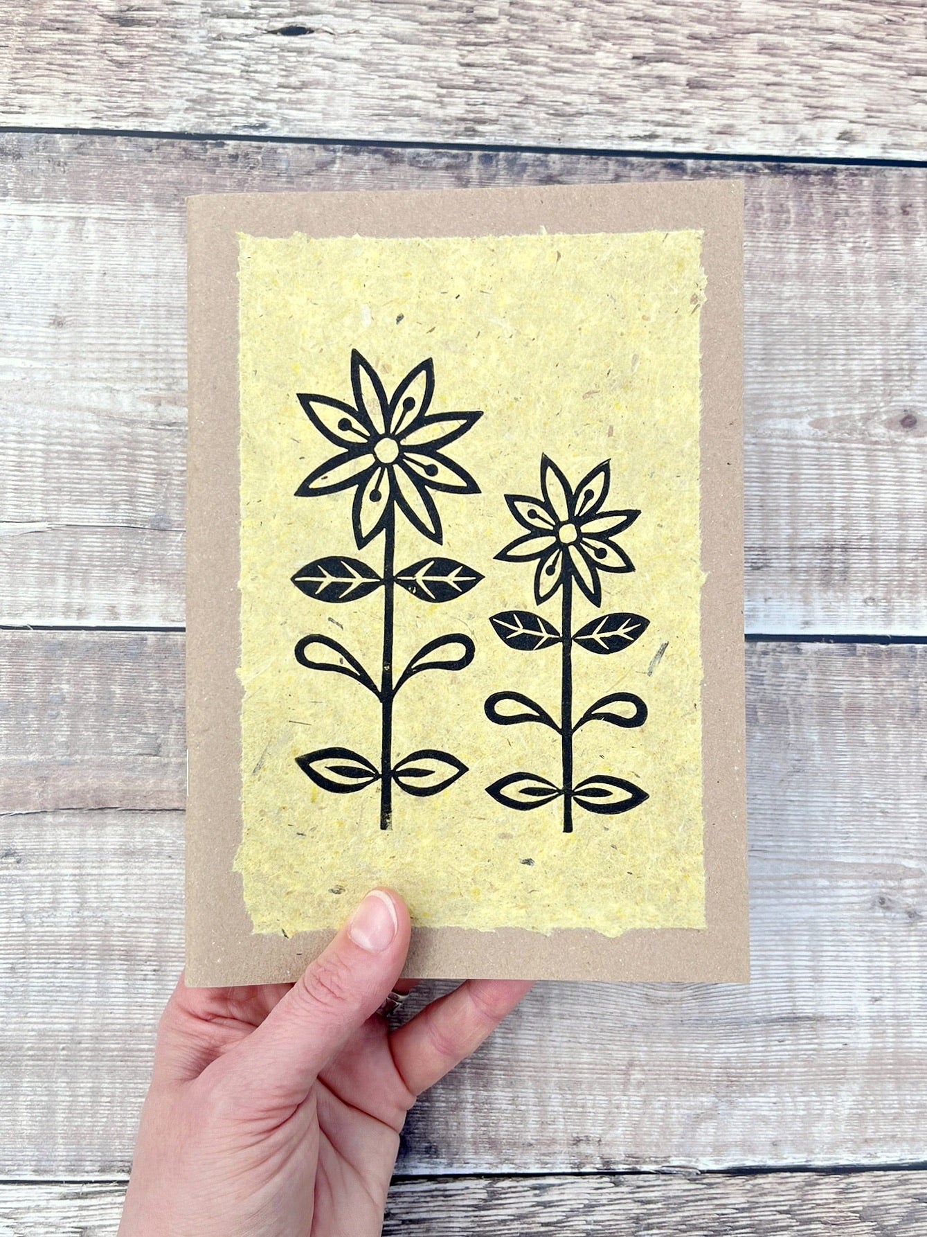 A5 kraft sketchbook with an original lino print of daisies on handmade yellow paper. Held in a hand against a wooden background.