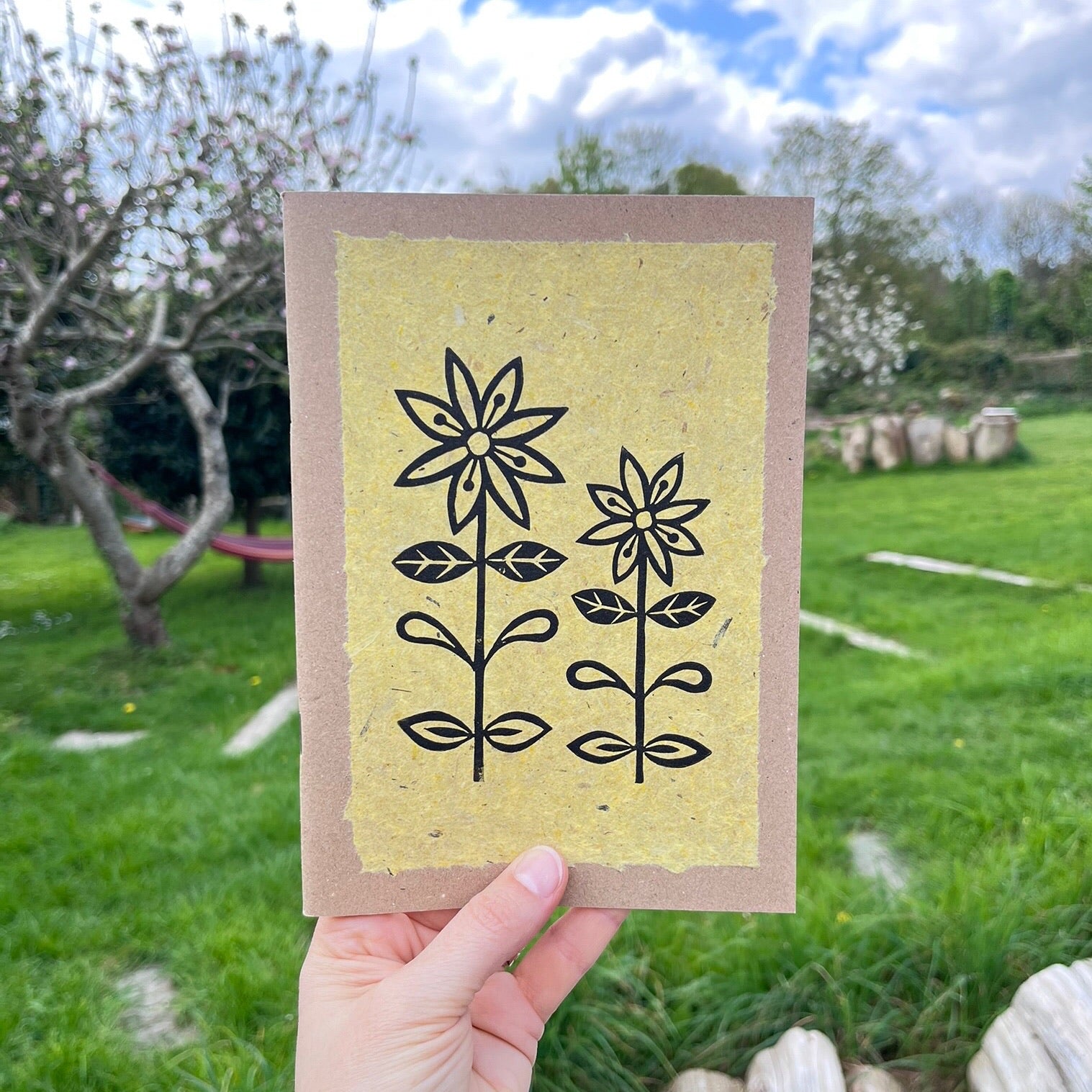A5 kraft sketchbook with an original lino print of daisies on handmade yellow paper. Held in a hand in a garden.