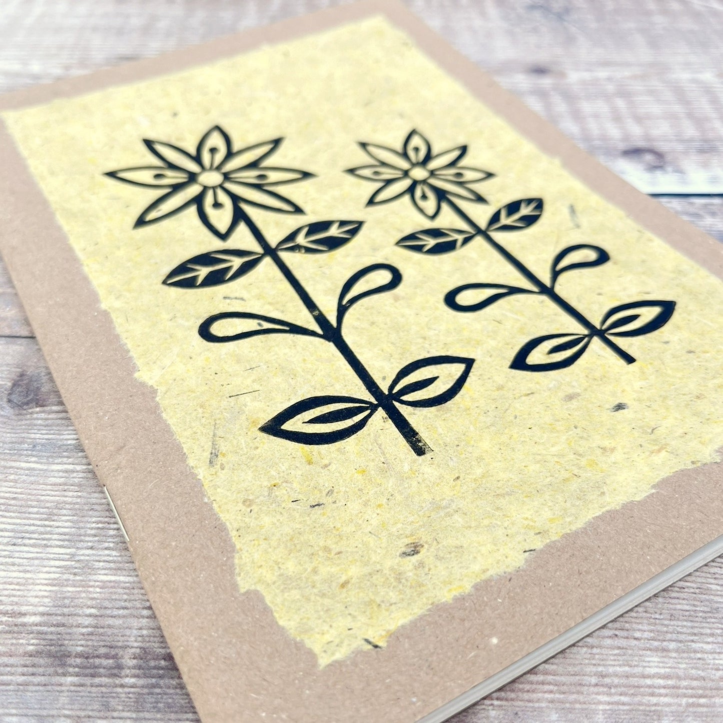 Close up of A5 kraft sketchbook with an original lino print of daisies on handmade yellow paper. Laid on a wooden background.