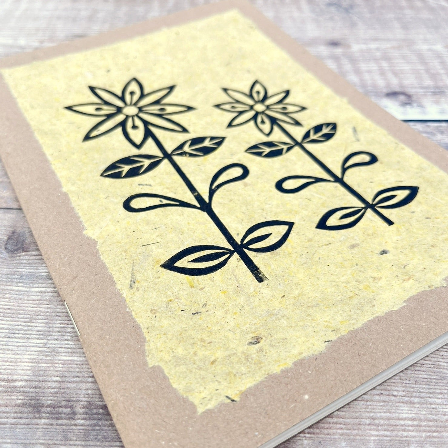 Close up of A5 kraft sketchbook with an original lino print of daisies on handmade yellow paper. Laid on a wooden background.