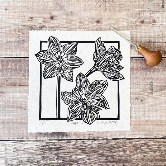 Lino print of Daffodils in black ink on off white paper, laid on a wooden surface. Photo includes lino cutting tool.