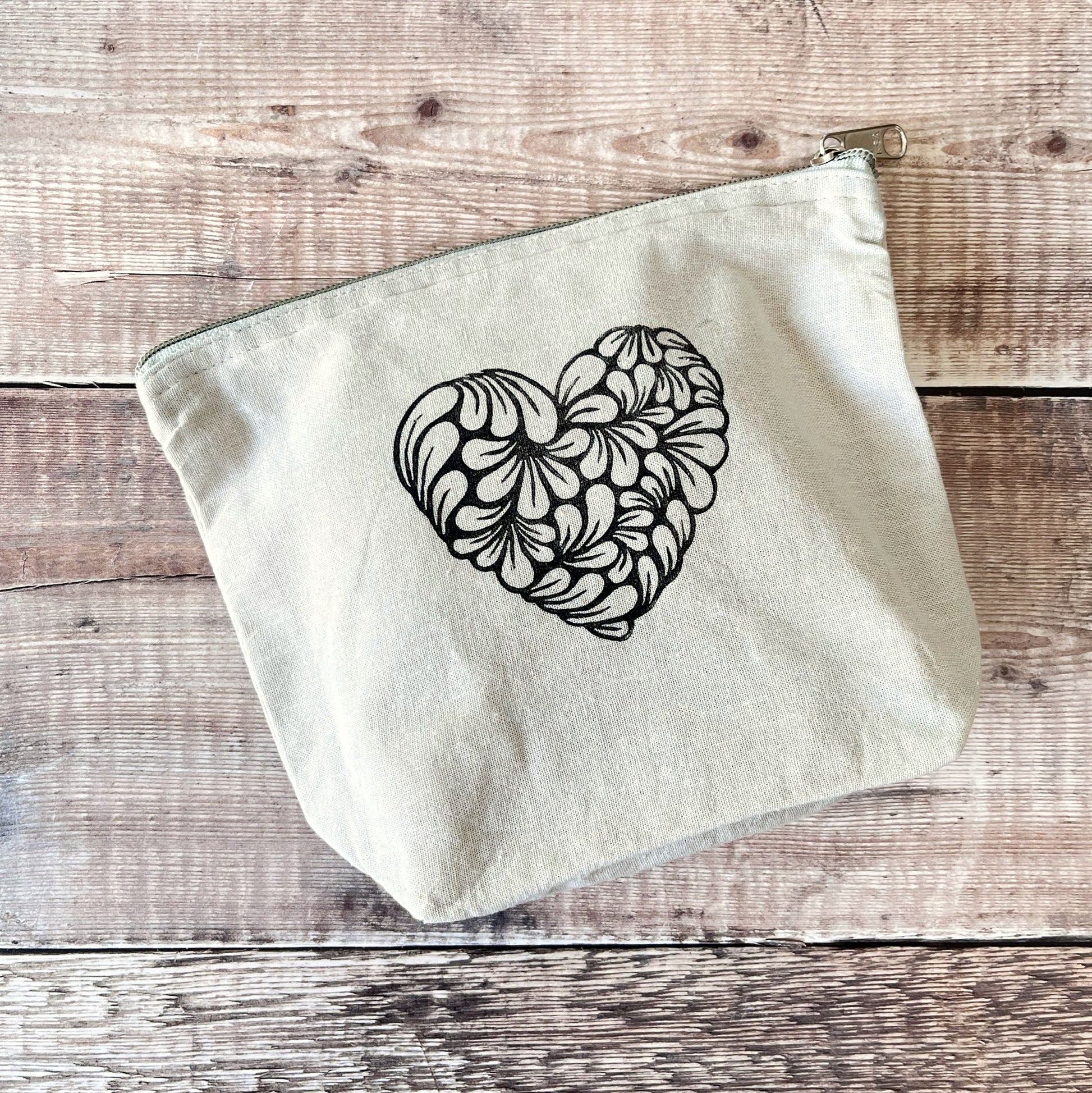 Pale grey toiletry bag with lino printed heart design. Laid on a wooden surface.