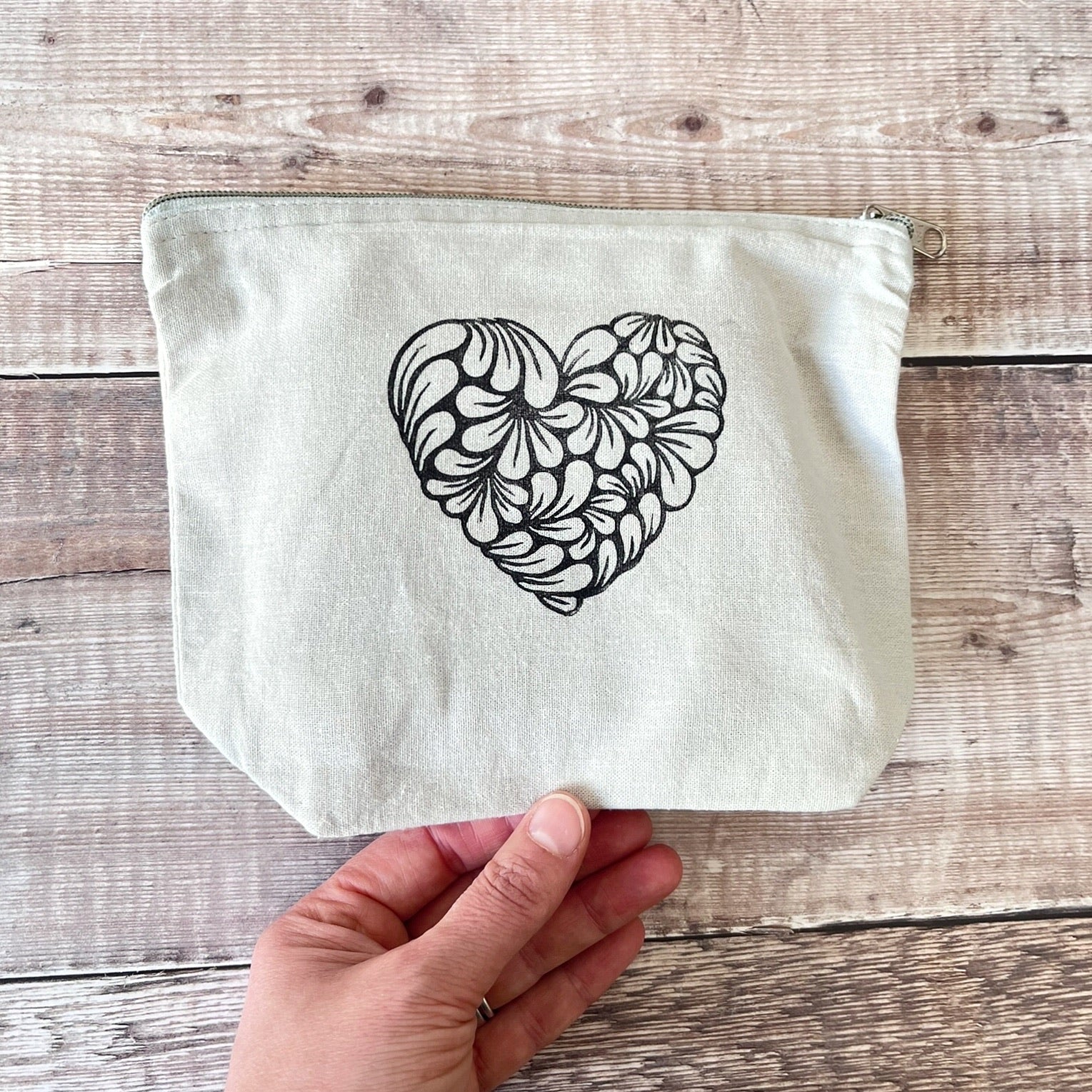 Pale grey toiletry bag with lino printed heart design. Held in a hand against a wooden surface.