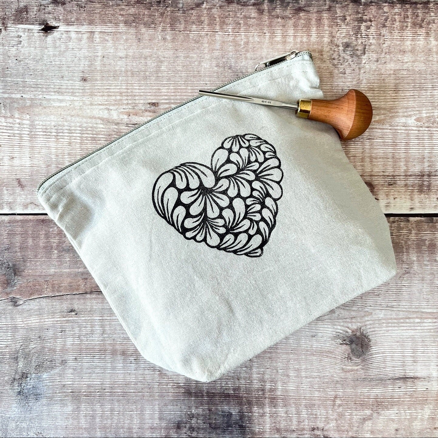 Pale grey toiletry bag with lino printed heart design. Laid on a wooden surface the photo includes the lino cutting tool used.
