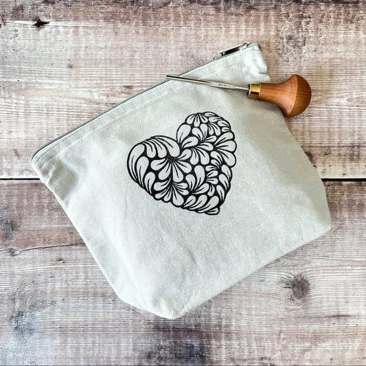 Pale grey toiletry bag with lino printed heart design. Laid on a wooden surface the photo includes the lino cutting tool used.