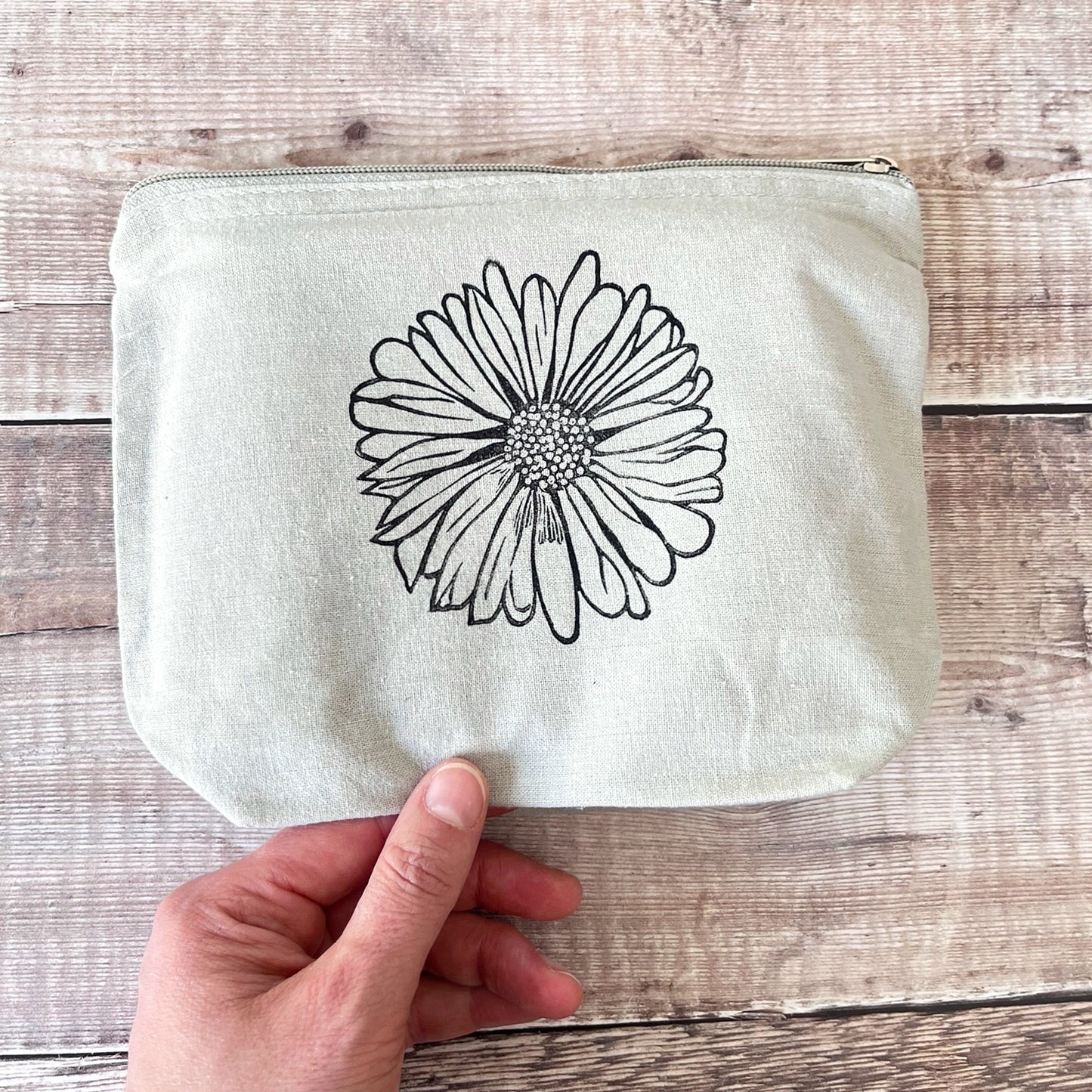 Pale grey cotton toiletry bag with a Daisy lino print held in a hand against a wooden surface. 