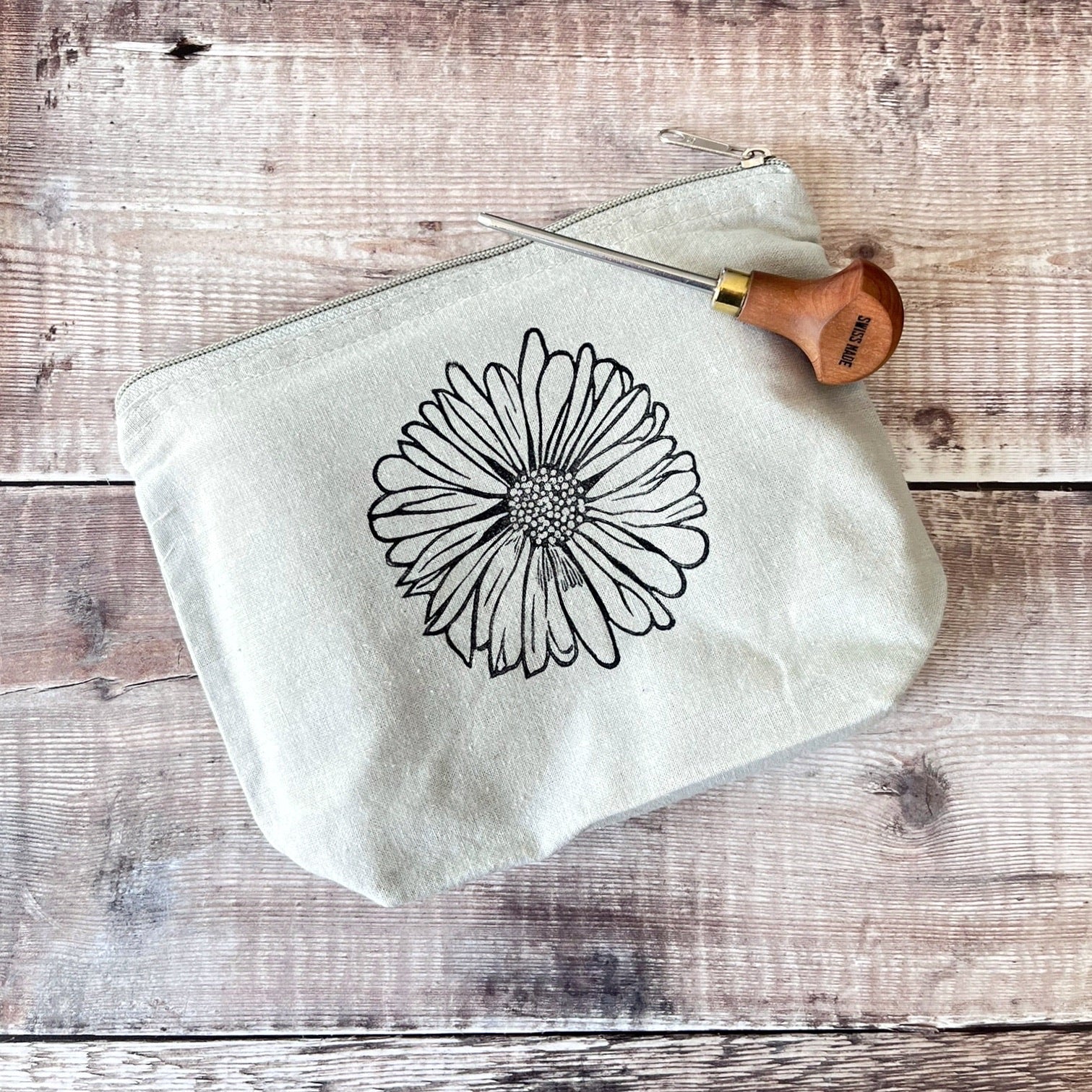 Pale grey cotton toiletry bag with a Daisy lino print laid on a wooden surface. Photo includes lino cutting tool.