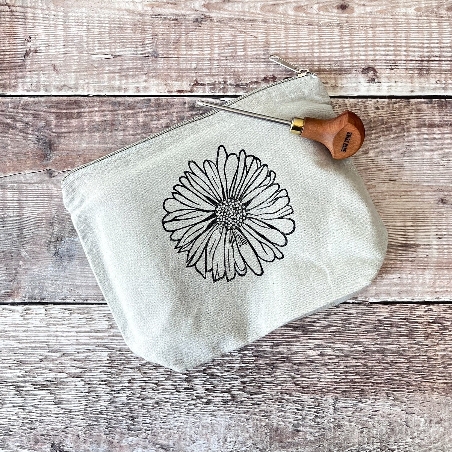 Pale grey cotton toiletry bag with a Daisy lino print laid on a wooden surface. Photo includes lino cutting tool.