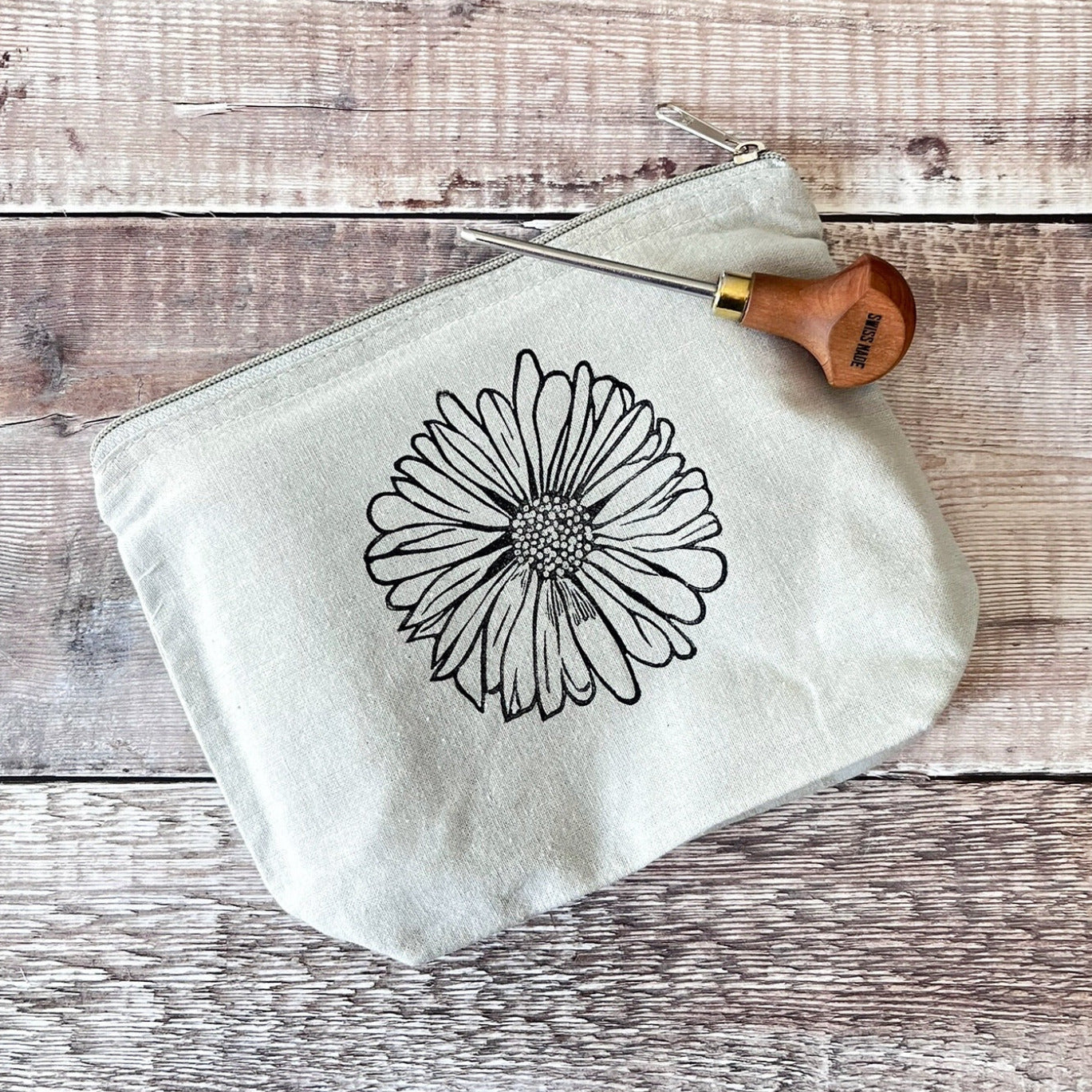 Pale grey cotton toiletry bag with a Daisy lino print laid on a wooden surface. Photo includes lino cutting tool.