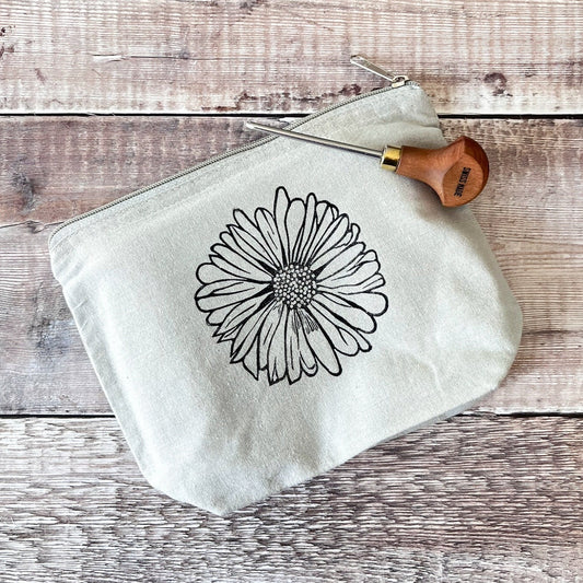 Pale grey cotton toiletry bag with a Daisy lino print laid on a wooden surface. Photo includes lino cutting tool.