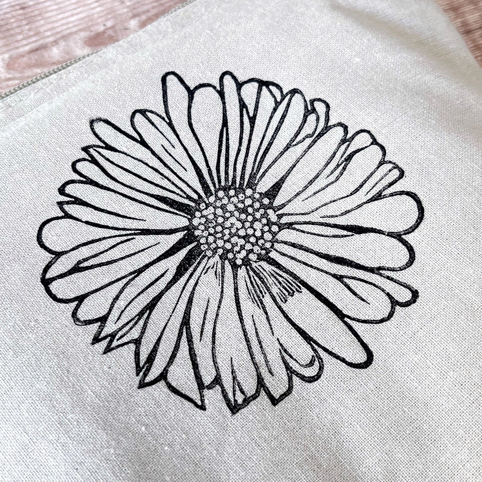 Close up of pale grey cotton toiletry bag with a Daisy lino print laid on a wooden surface. 