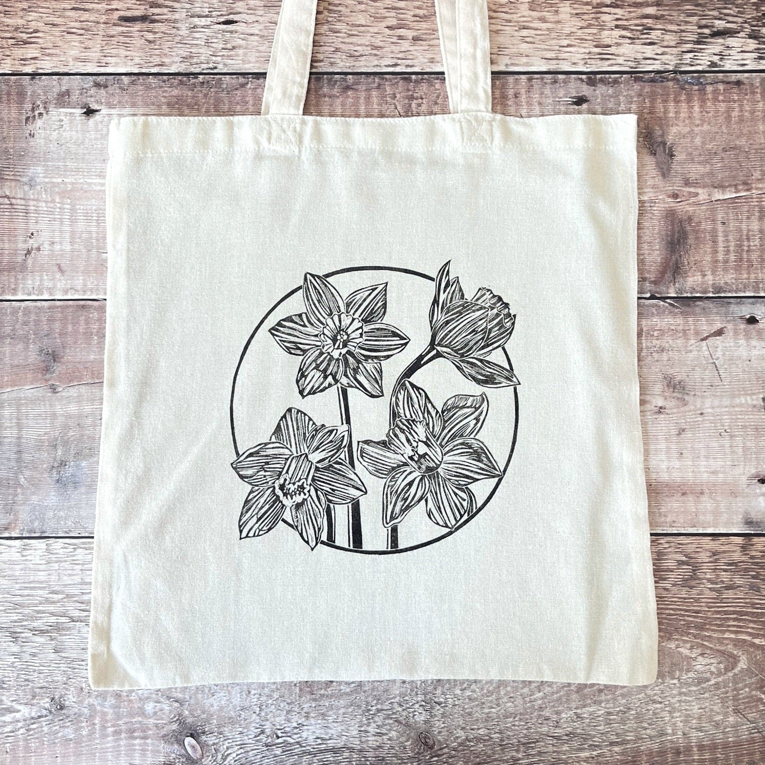 Cream cotton tote bag with a lino printed Daffodils laid on a wooden surface