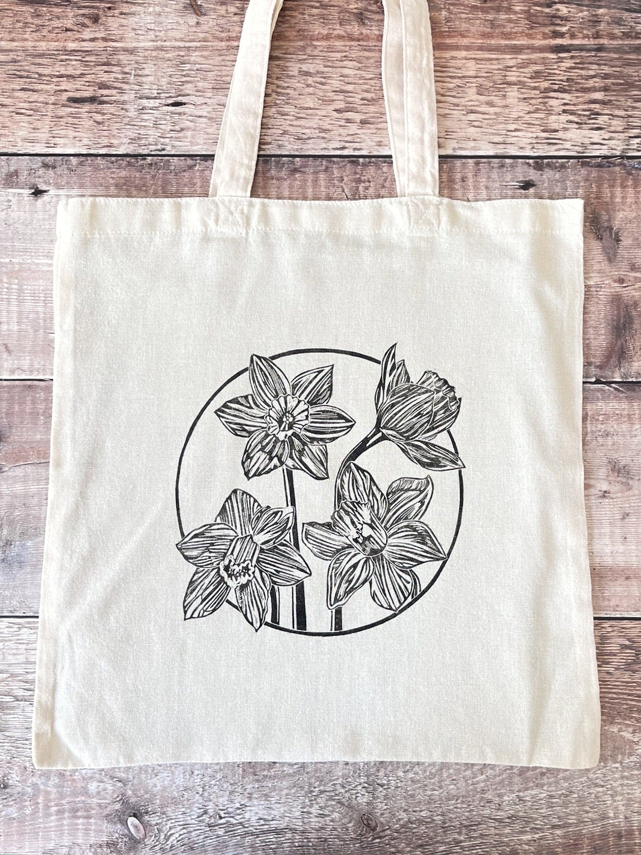 Cream cotton tote bag with a lino printed Daffodils laid on a wooden surface