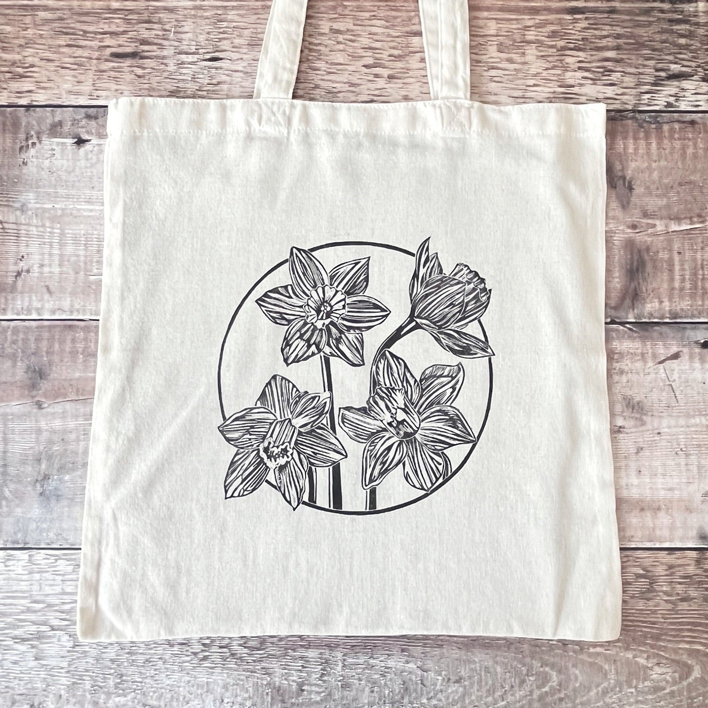 Cream cotton tote bag with a lino printed Daffodils laid on a wooden surface