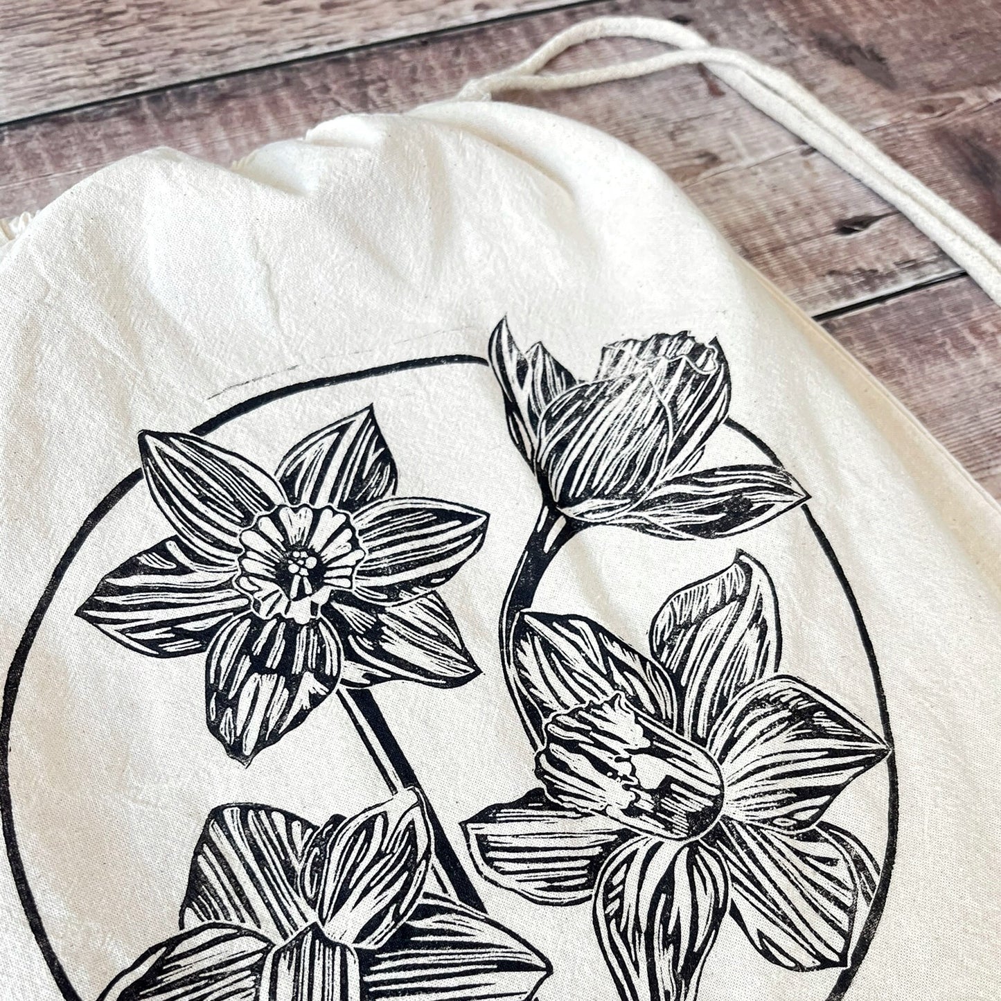Close up of lino print of Daffodils on a natural cotton drawstring bag laid on a wooden surface