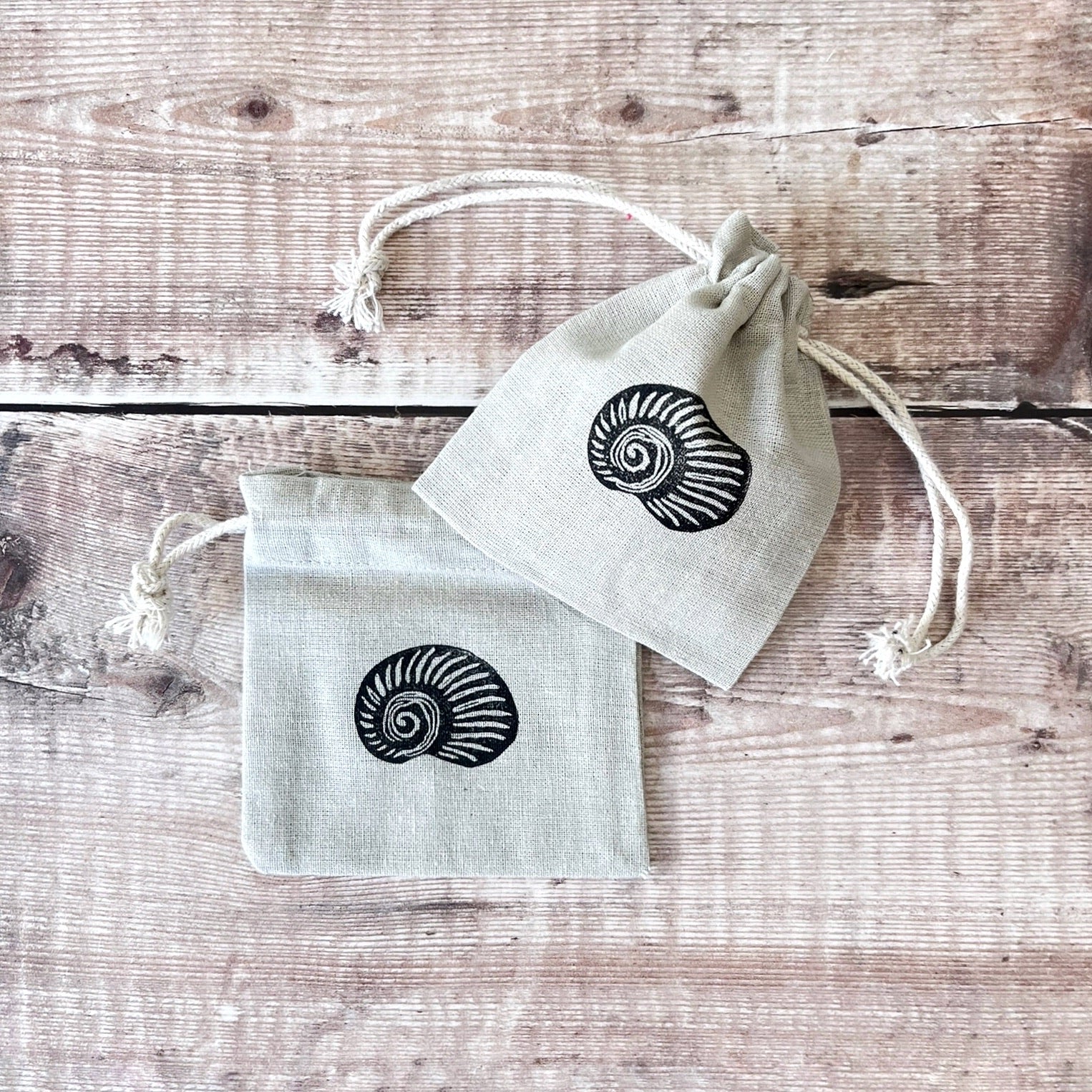 Two grey drawstring cotton gift bags with a lino printed shell, laid on a wooden surface.