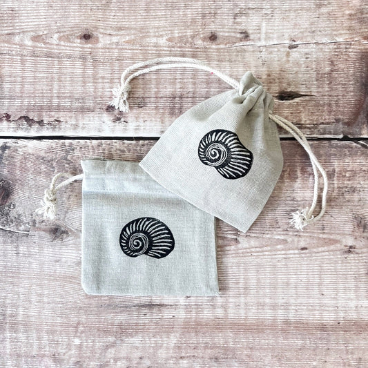 Two grey drawstring cotton gift bags with a lino printed shell, laid on a wooden surface.