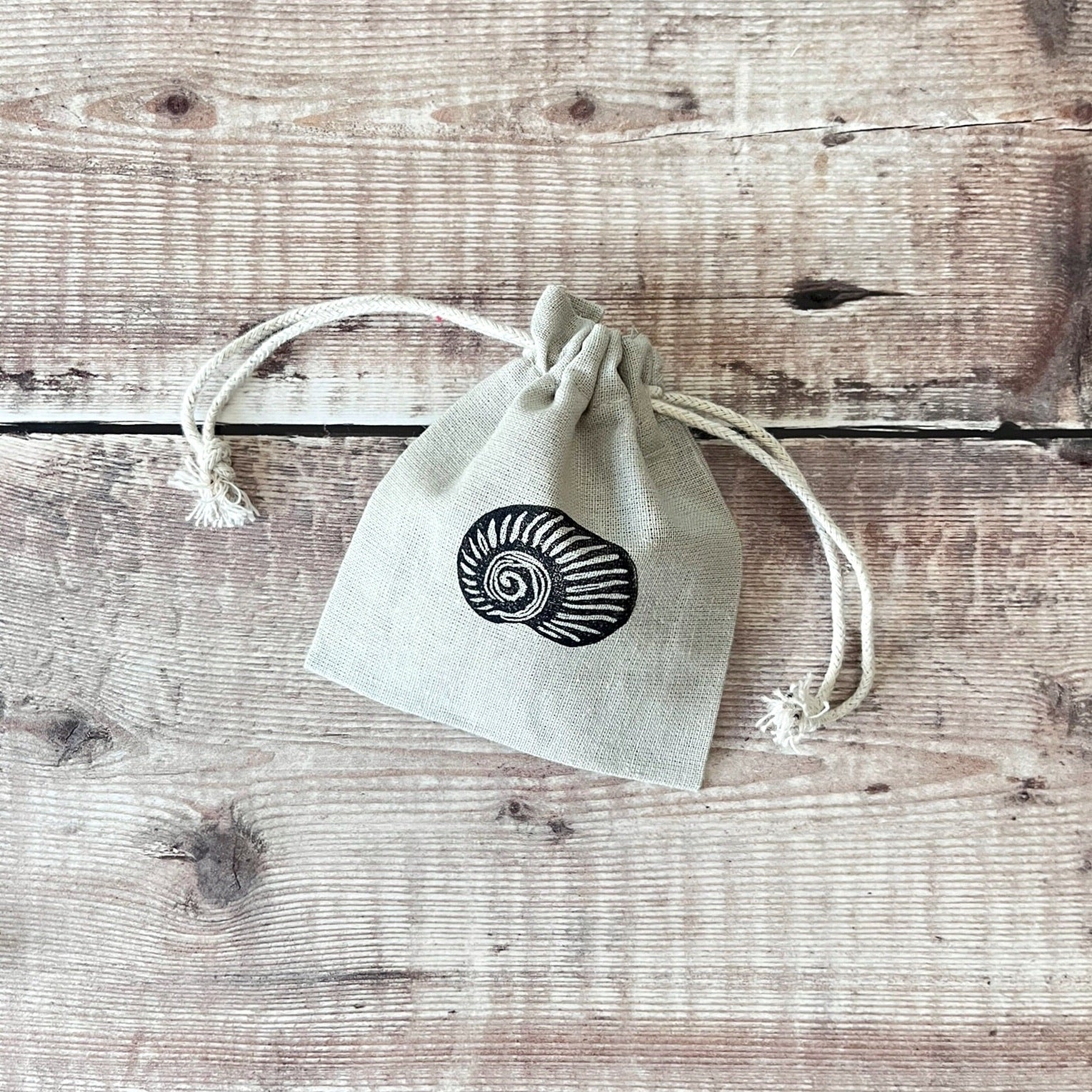 Grey drawstring cotton gift bag with a lino printed shell, laid on a wooden surface.