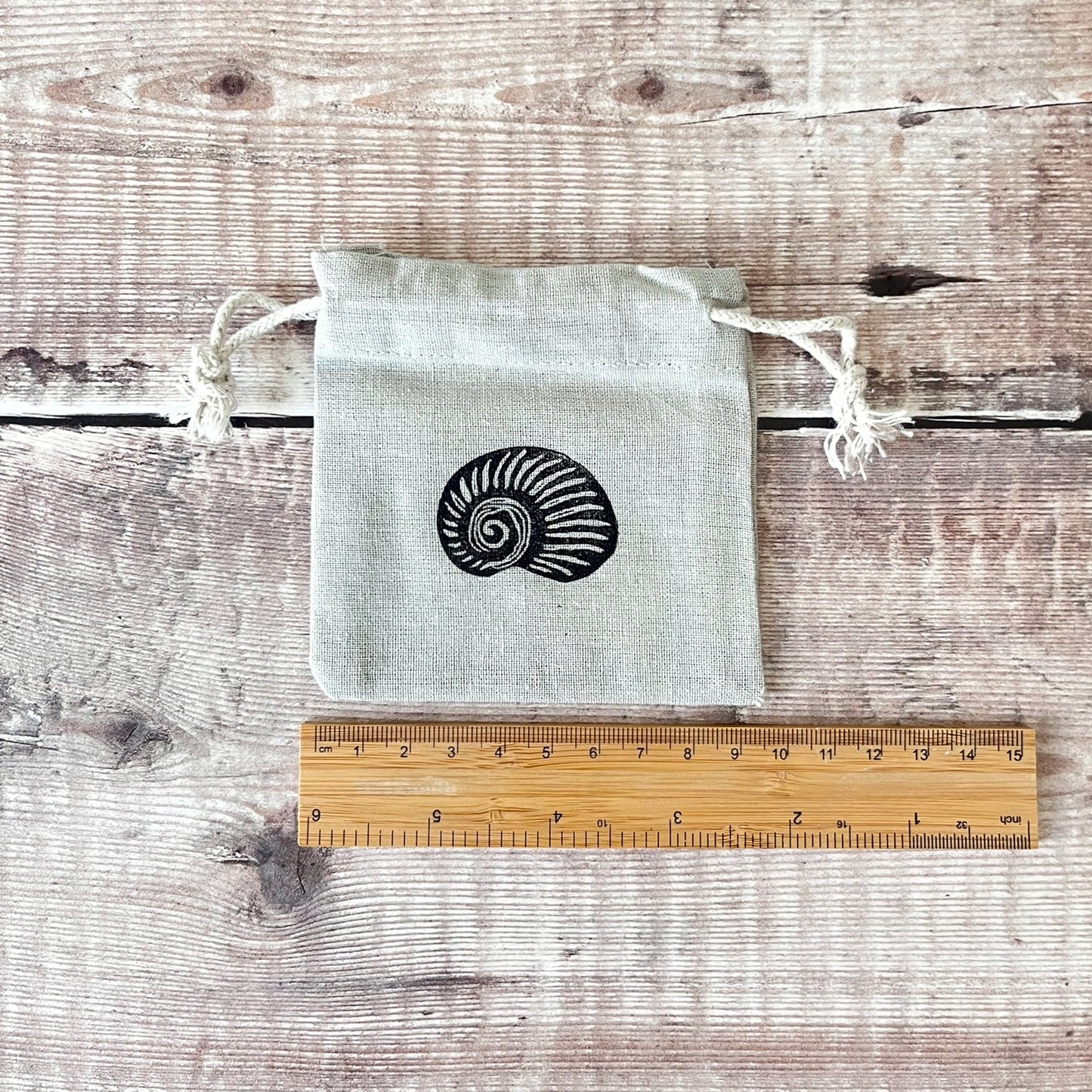 Grey drawstring cotton gift bag with a lino printed shell, laid flat next to a wooden ruler on a wooden surface.