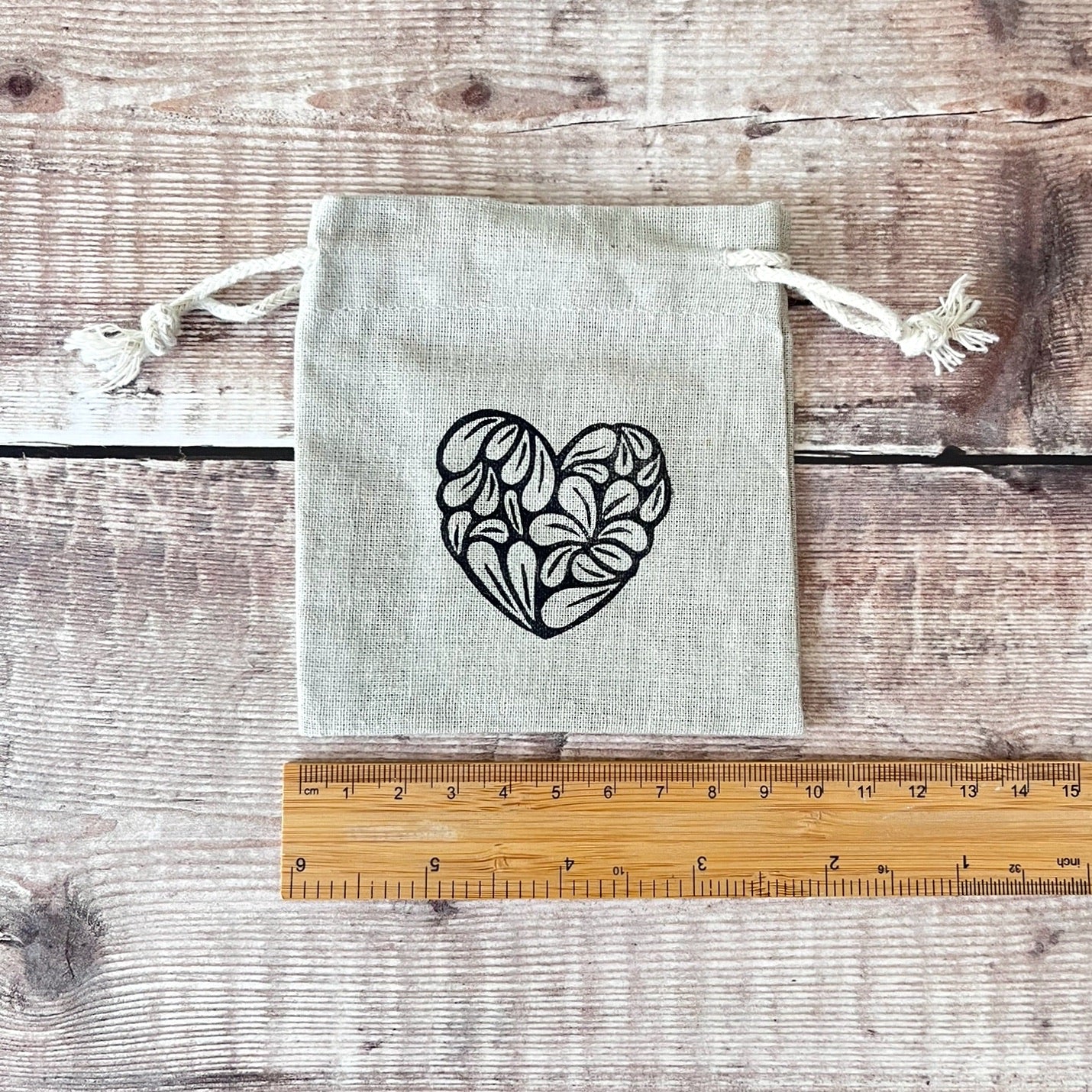 Light gray drawstring bag with a black heart design on a wooden surface with a ruler for scale.