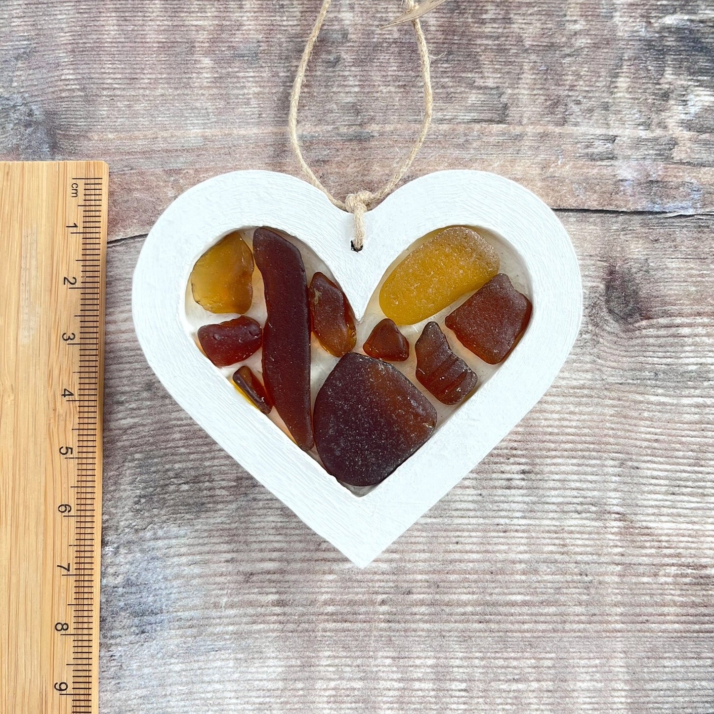 Heart-shaped sea glass hanging decoration next to a ruler for scale and laid on a wooden surface