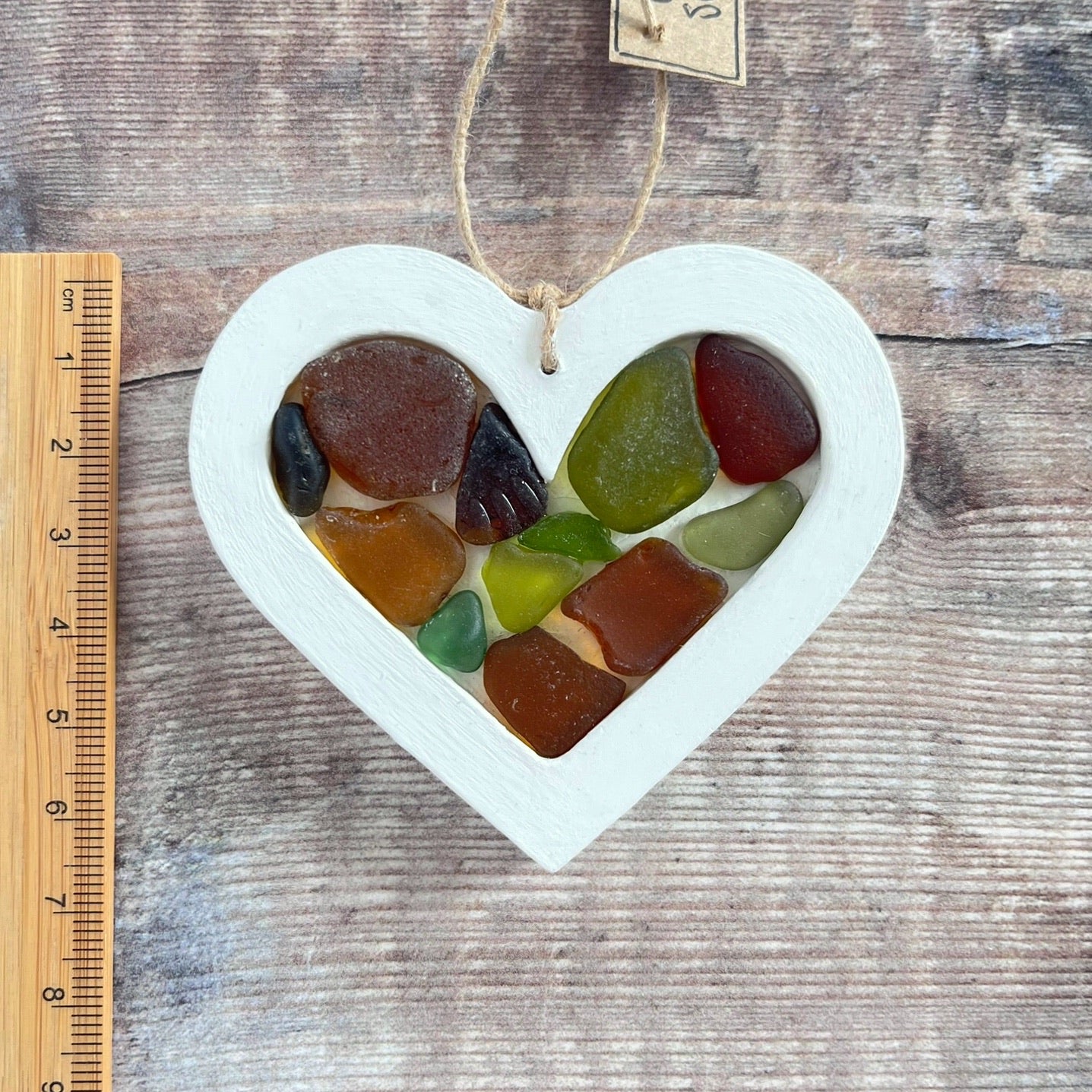 Heart-shaped sea glass hanging decoration next to a ruler for scale laid on a wooden surface
