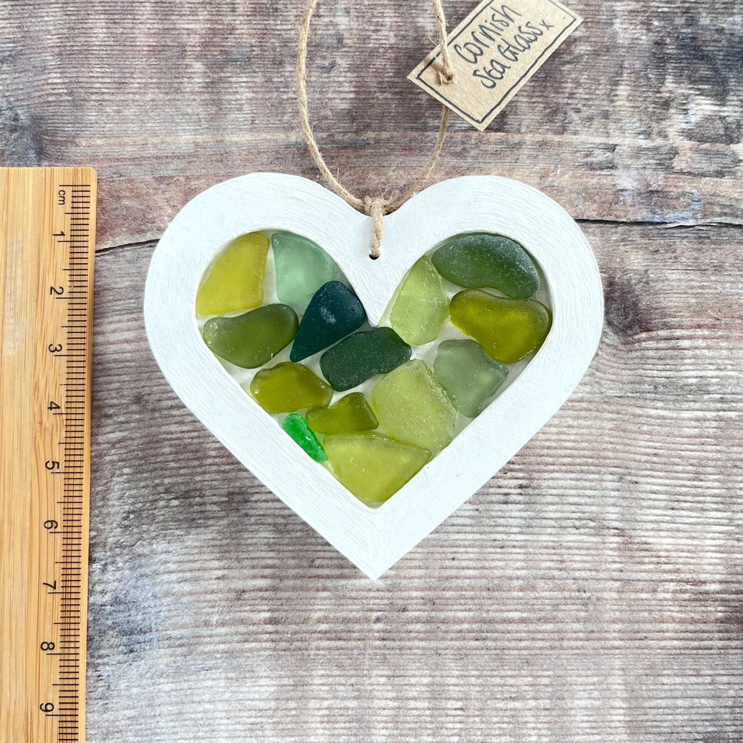 Heart-shaped sea glass hanging decoration next to a ruler for scale and laid on a wooden surface