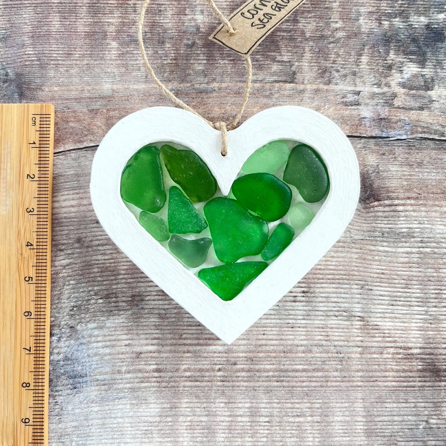 Heart-shaped sea glass hanging decoration next to a ruler for scale and laid on a wooden surface