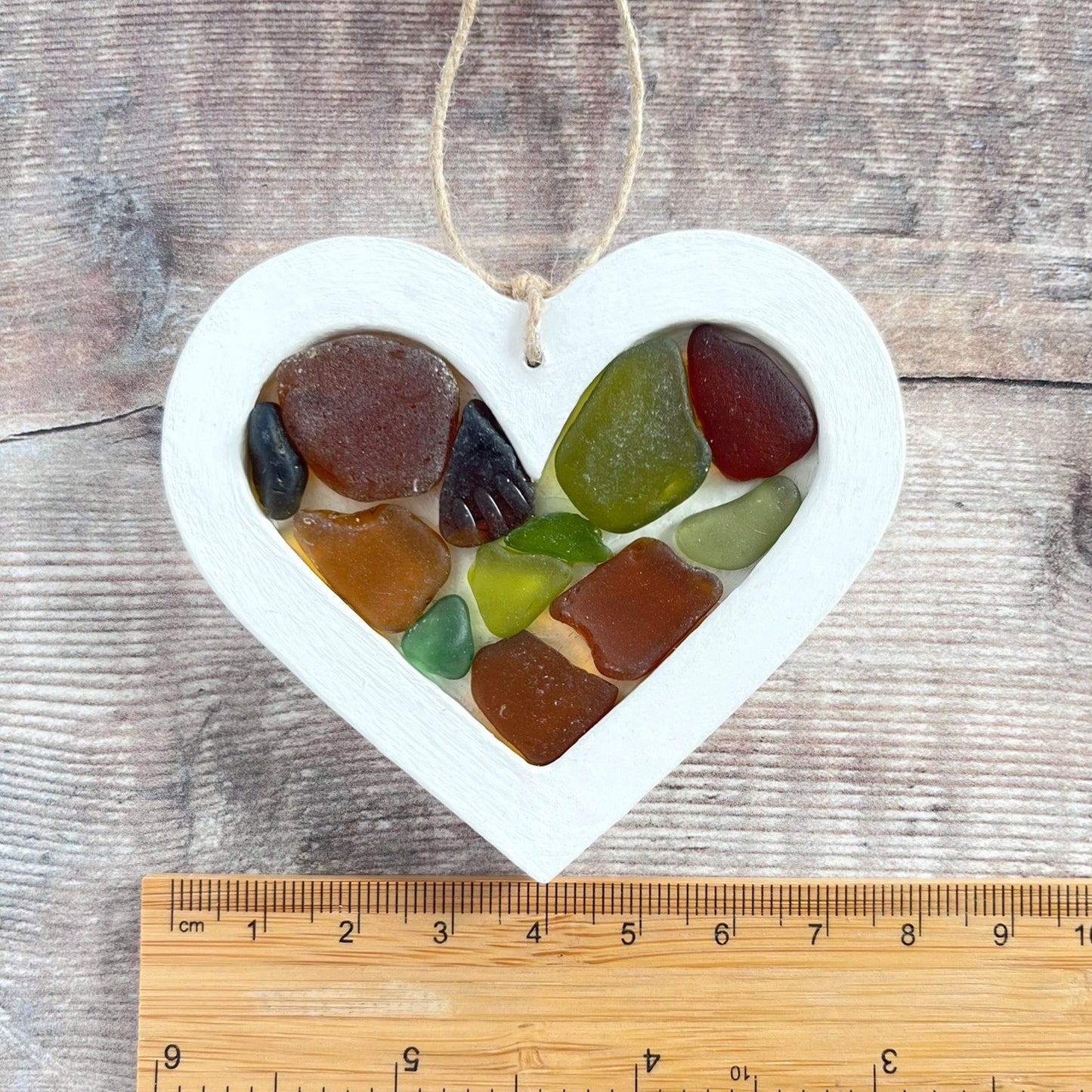 Heart-shaped sea glass hanging decoration next to a ruler for scale laid on a wooden surface