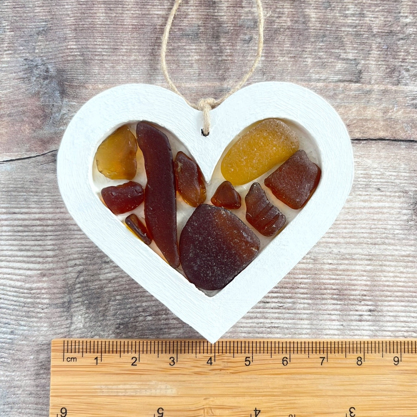 Heart-shaped sea glass hanging decoration next to a ruler for scale and laid on a wooden surface