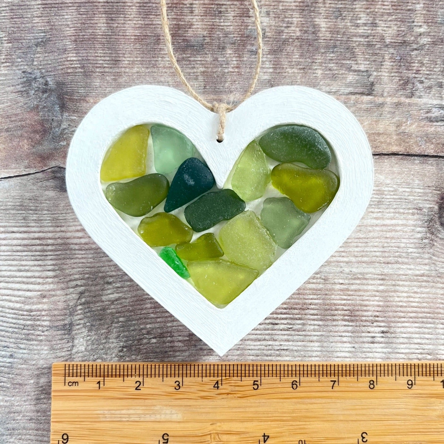 Heart-shaped sea glass hanging decoration next to a ruler for scale laid on a wooden surface