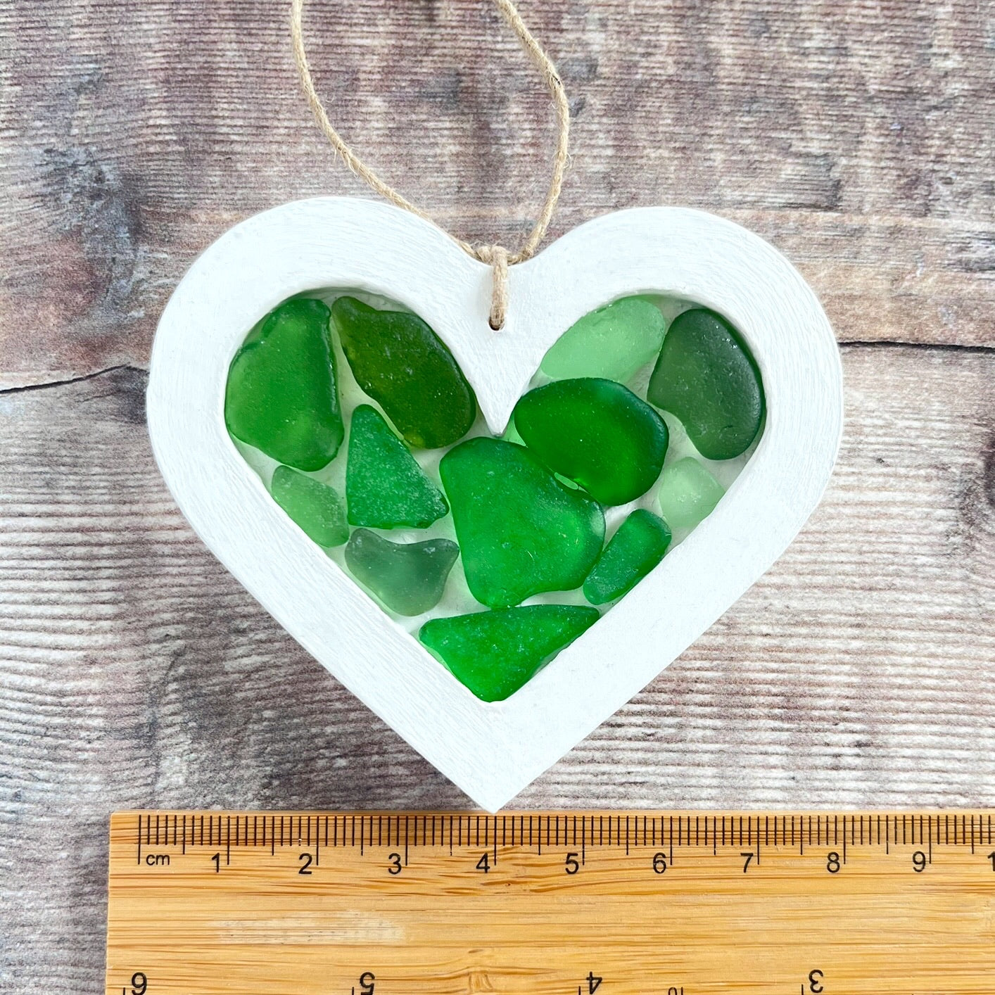 Heart-shaped sea glass hanging decoration next to a ruler for scale and laid on a wooden surface