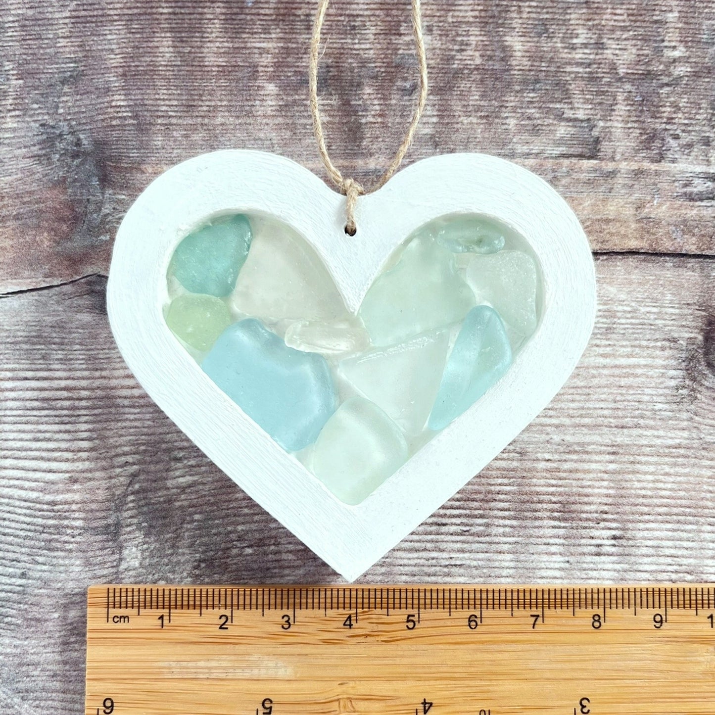 Heart-shaped sea glass hanging decoration next to a ruler for scale and laid on a wooden surface