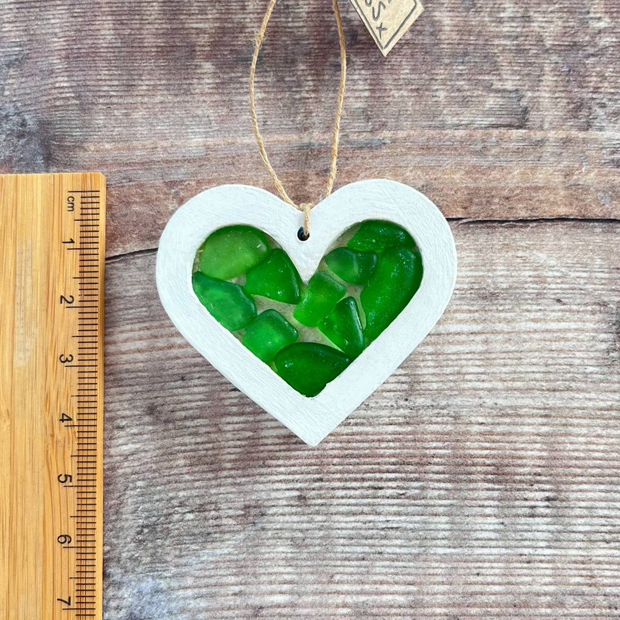 Heart-shaped sea glass hanging decoration next to a ruler for scale and laid on a wooden surface