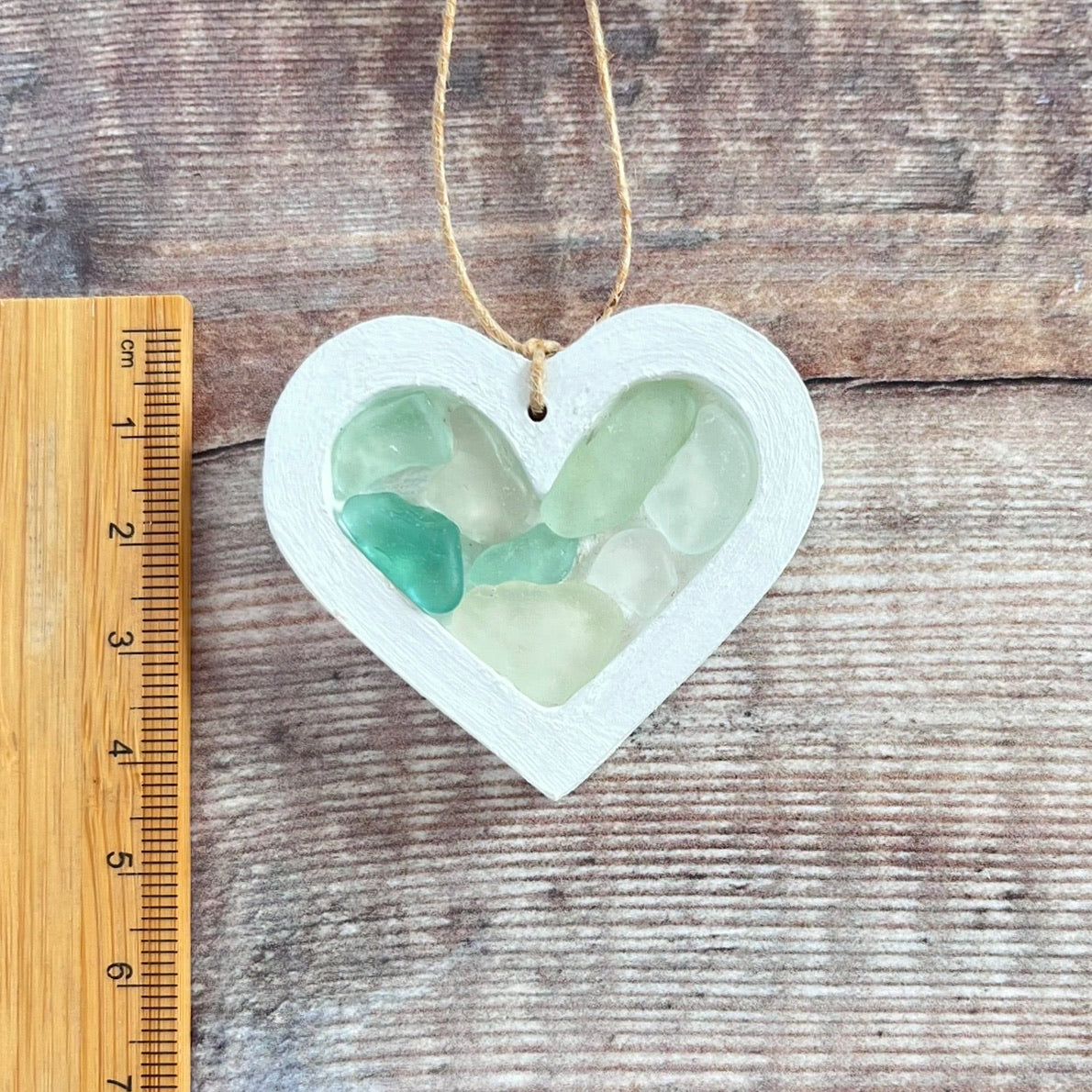 Heart-shaped sea glass hanging decoration next to a ruler for scale and laid on a wooden surface