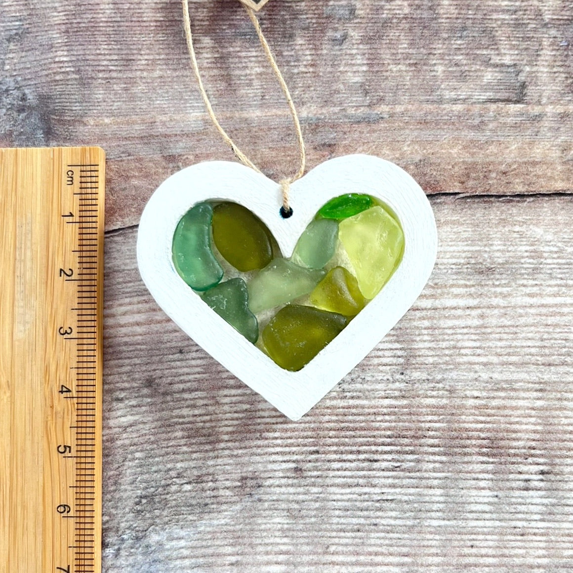 Heart-shaped sea glass hanging decoration next to a ruler for scale and laid on a wooden surface