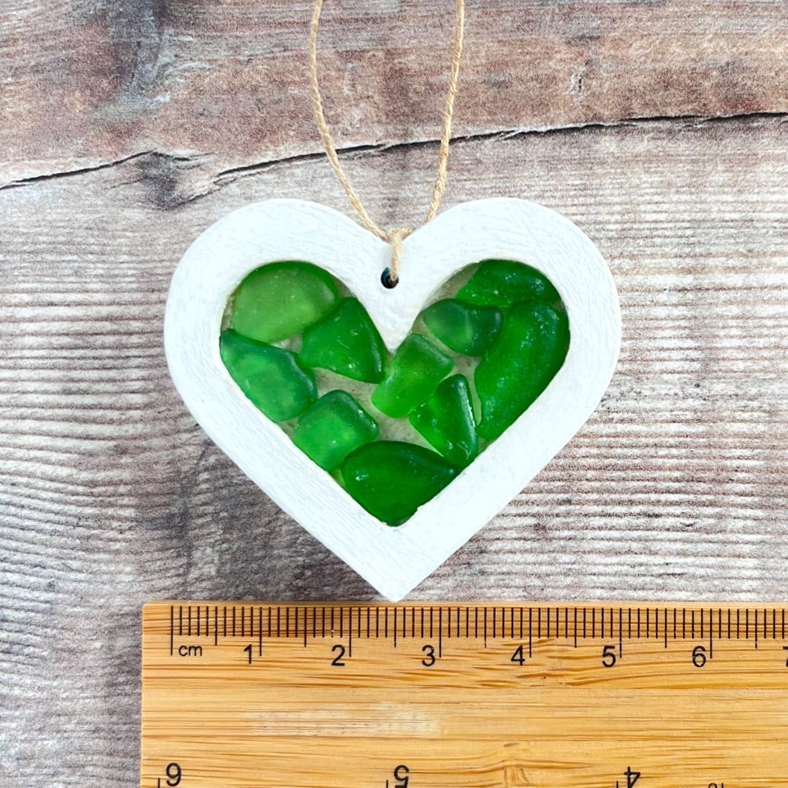 Heart-shaped sea glass hanging decoration next to a ruler for scale and laid on a wooden surface