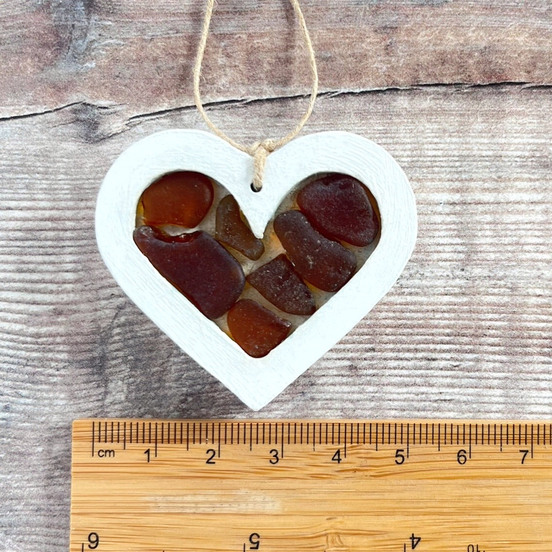 Heart-shaped sea glass hanging decoration next to a ruler for scale and laid on a wooden surface