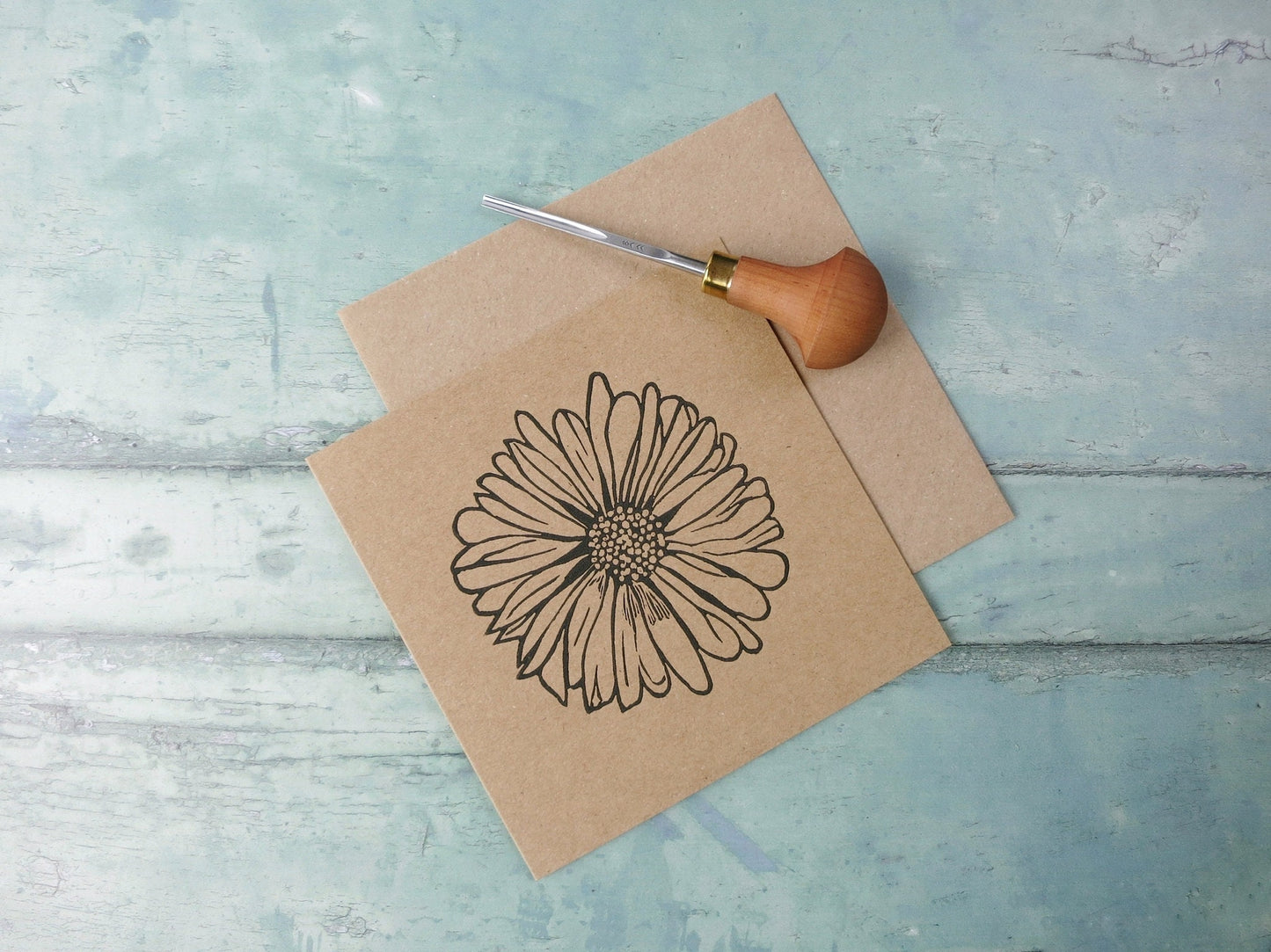 Kraft paper card with an original daisy lino print laid on a textured surface. Photo includes lino cutting tool