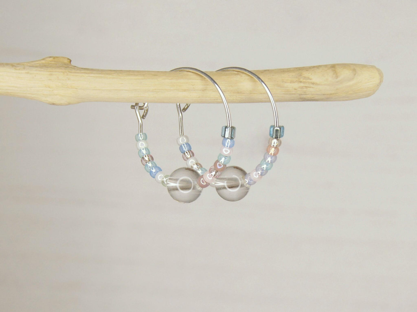 Pair of pastel seed bead and clear crystal hoop earrings hanging on driftwood against a pale background